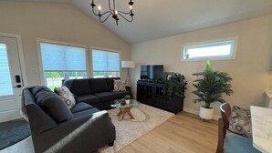 A spacious living room with a dark grey sofa, a glass coffee table, a black entertainment unit, and a potted plant. - Duplexes on Hawthorne (Dauphin)