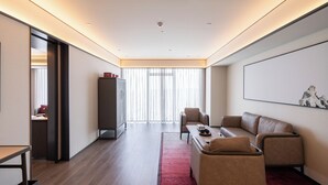 A modern hotel room with a sofa, armchairs, a coffee table, and a large window with sheer curtains.