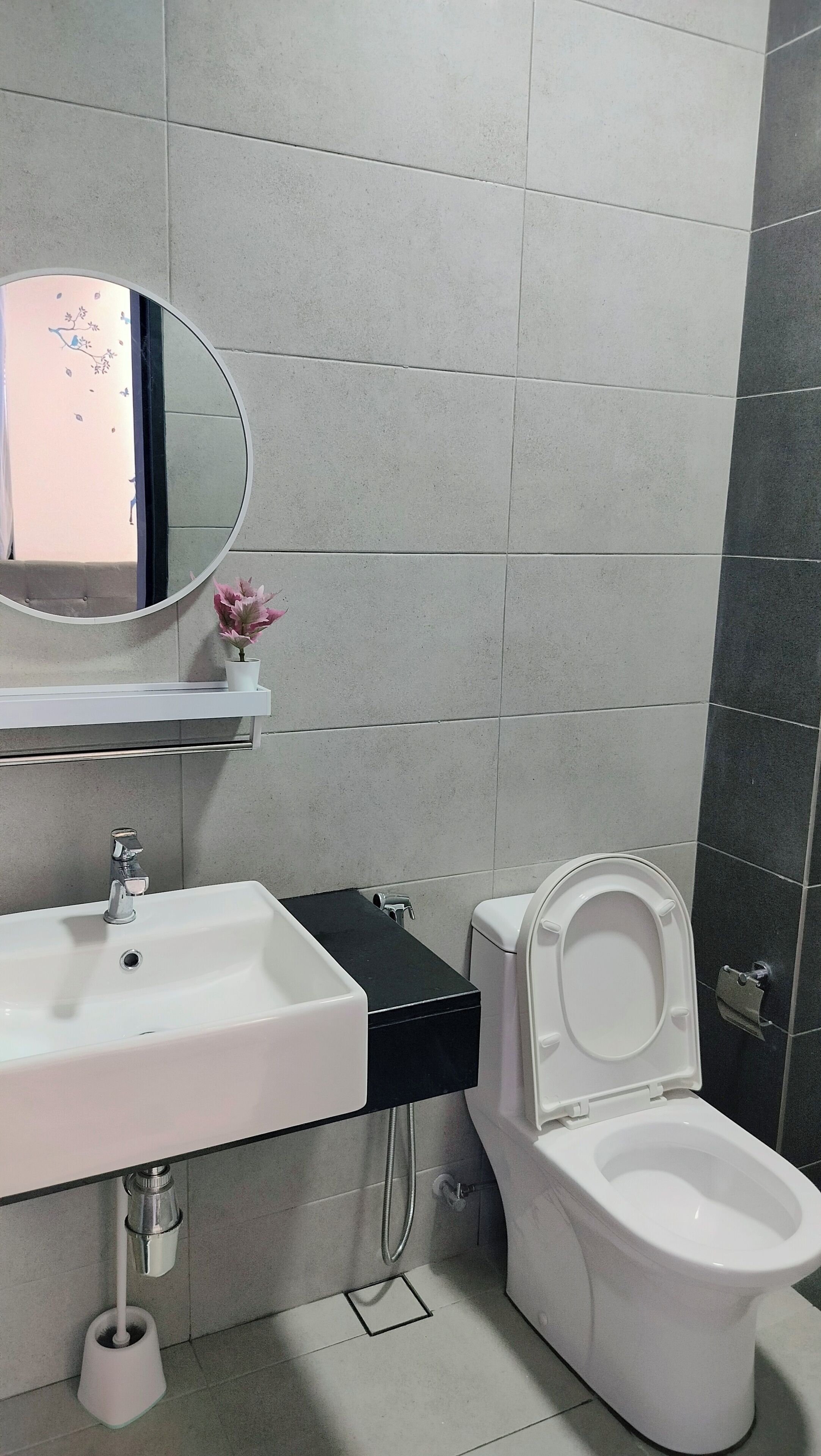 Et moderne badeværelse med en hvid vask, et rundt spejl, et toilet og en lille hylde med en potteplante.