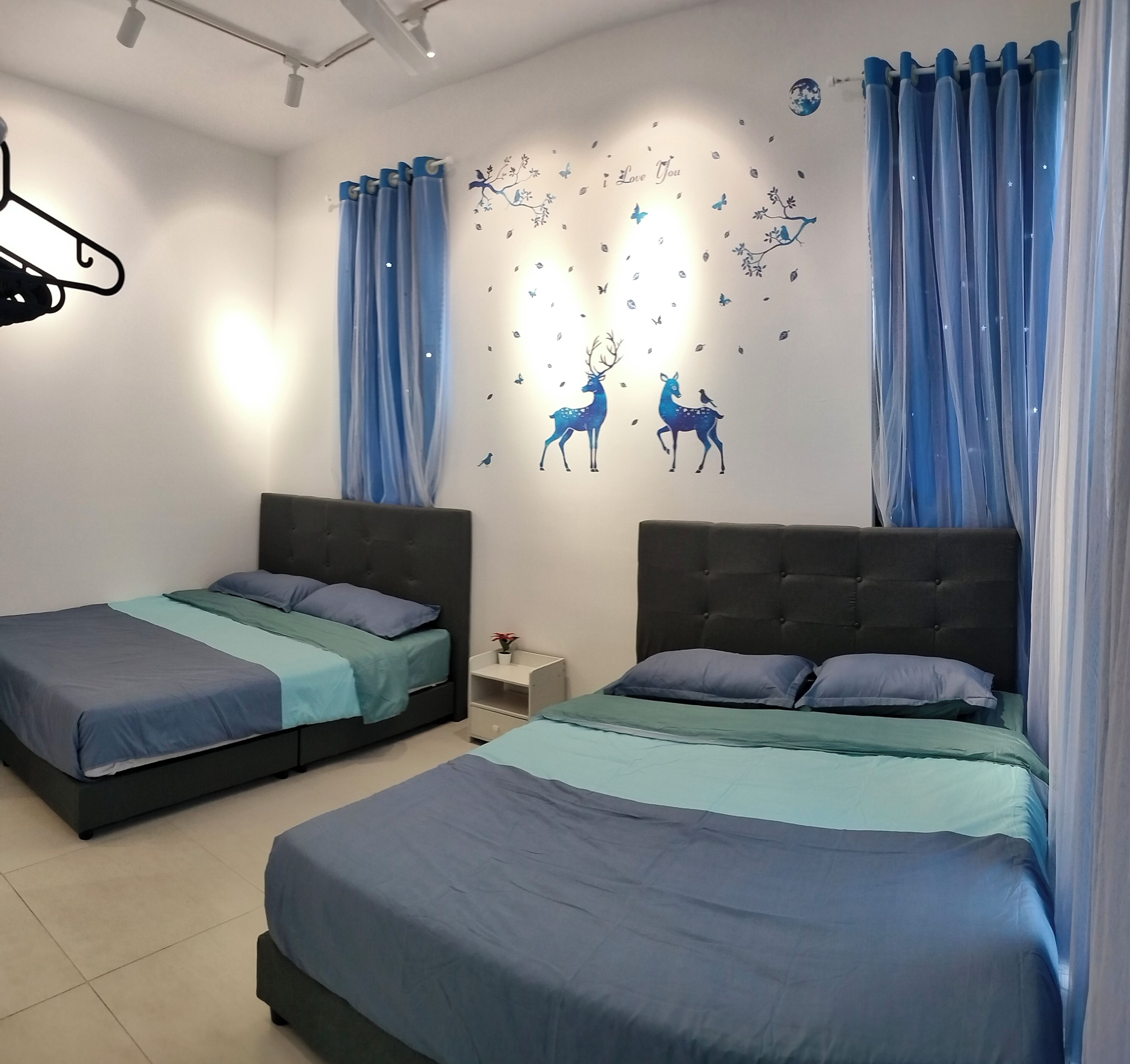 A bedroom with two beds, each with a dark headboard and light blue bedding. There is a wall decoration of deer and butterflies, and blue curtains on the windows.