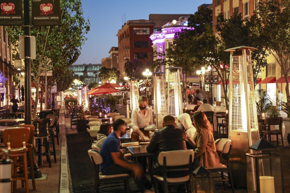 Un área de comedor al aire libre con mesas y sillas, rodeada de edificios y árboles.