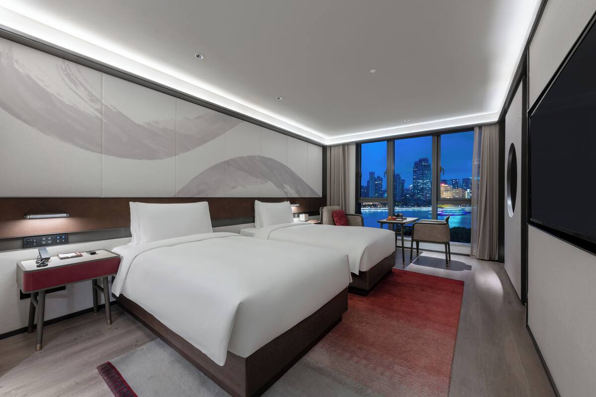 A modern hotel room with two beds, a desk, and a large window offering a city view.