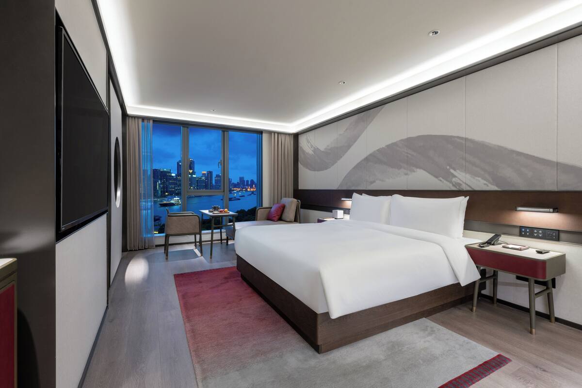 A modern hotel room with a large bed, a desk, and a chair. There is a city view through the window.