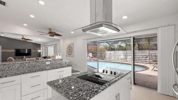Una cocina moderna con encimera de granito, armarios empotrados, una isla central y un amplio ventanal con vistas a un jardín con piscina.