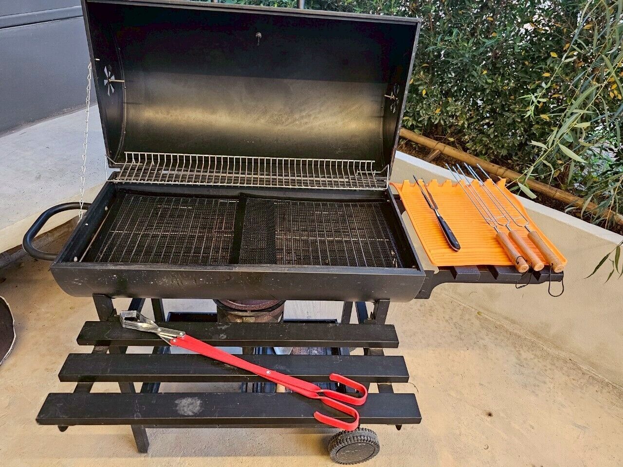Una parrilla portátil con rejilla metálica, una herramienta roja y brochetas de madera sobre un soporte de metal negro.