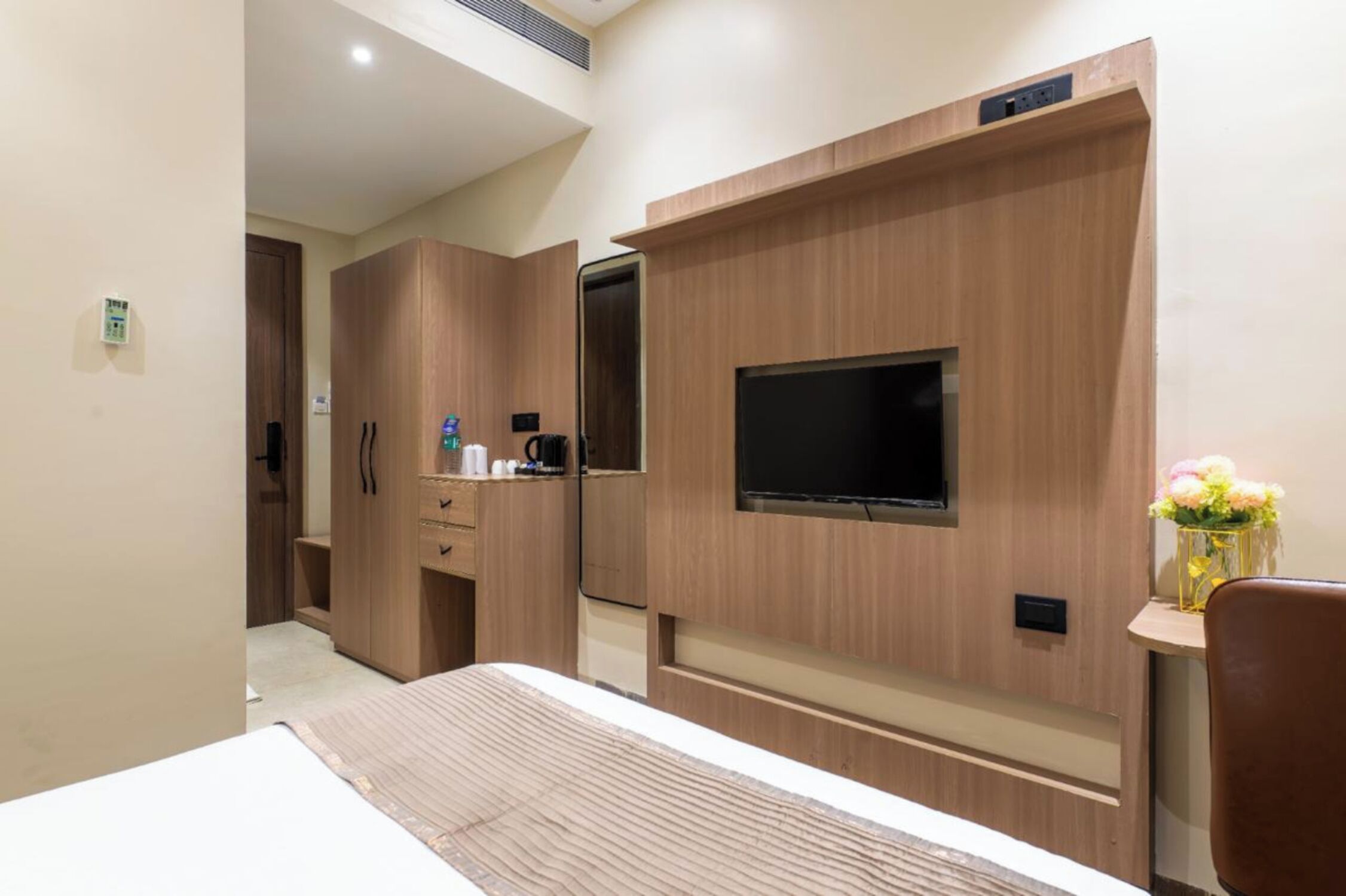 A hotel room with a large bed, wooden wardrobe, a flat-screen TV mounted on the wall, a small table with a vase of flowers, and a desk with a coffee maker.
