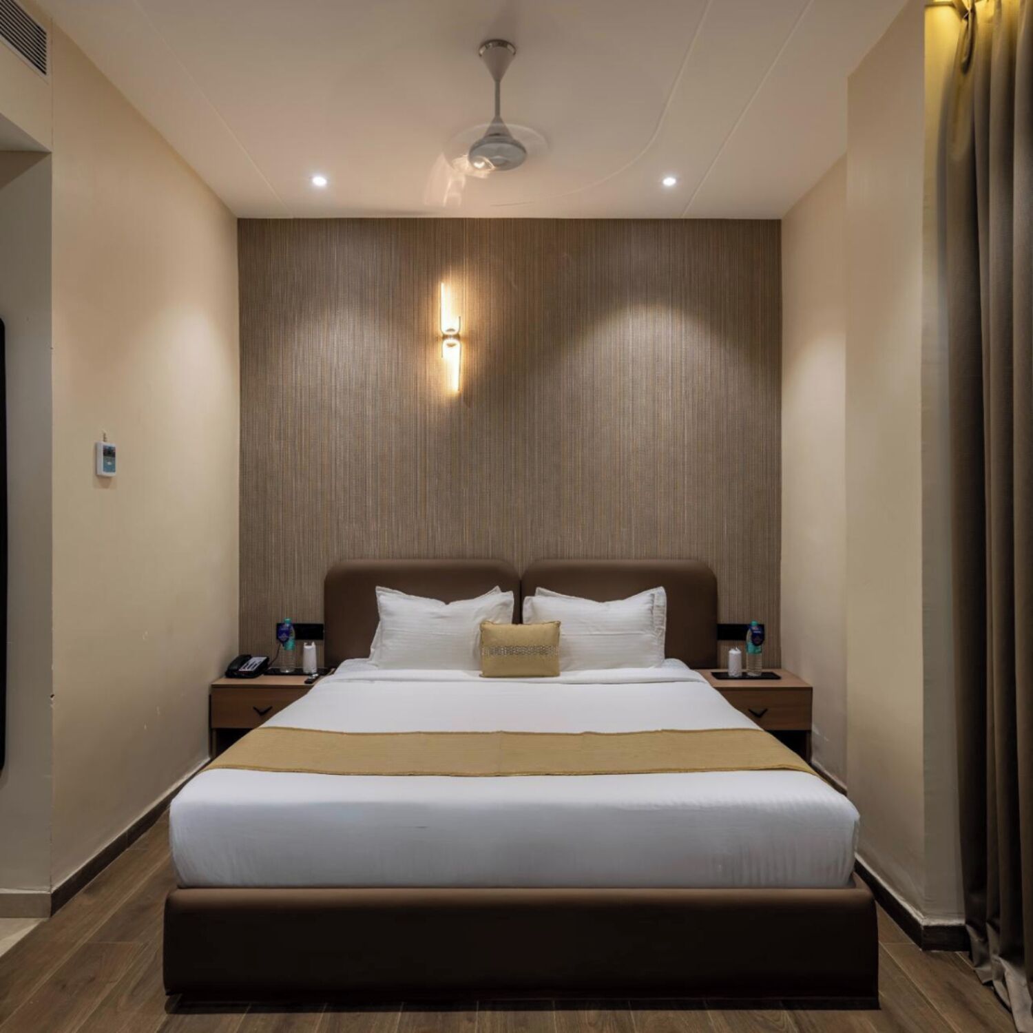 A modern hotel room with a large bed, wooden panel wall, and a ceiling fan.