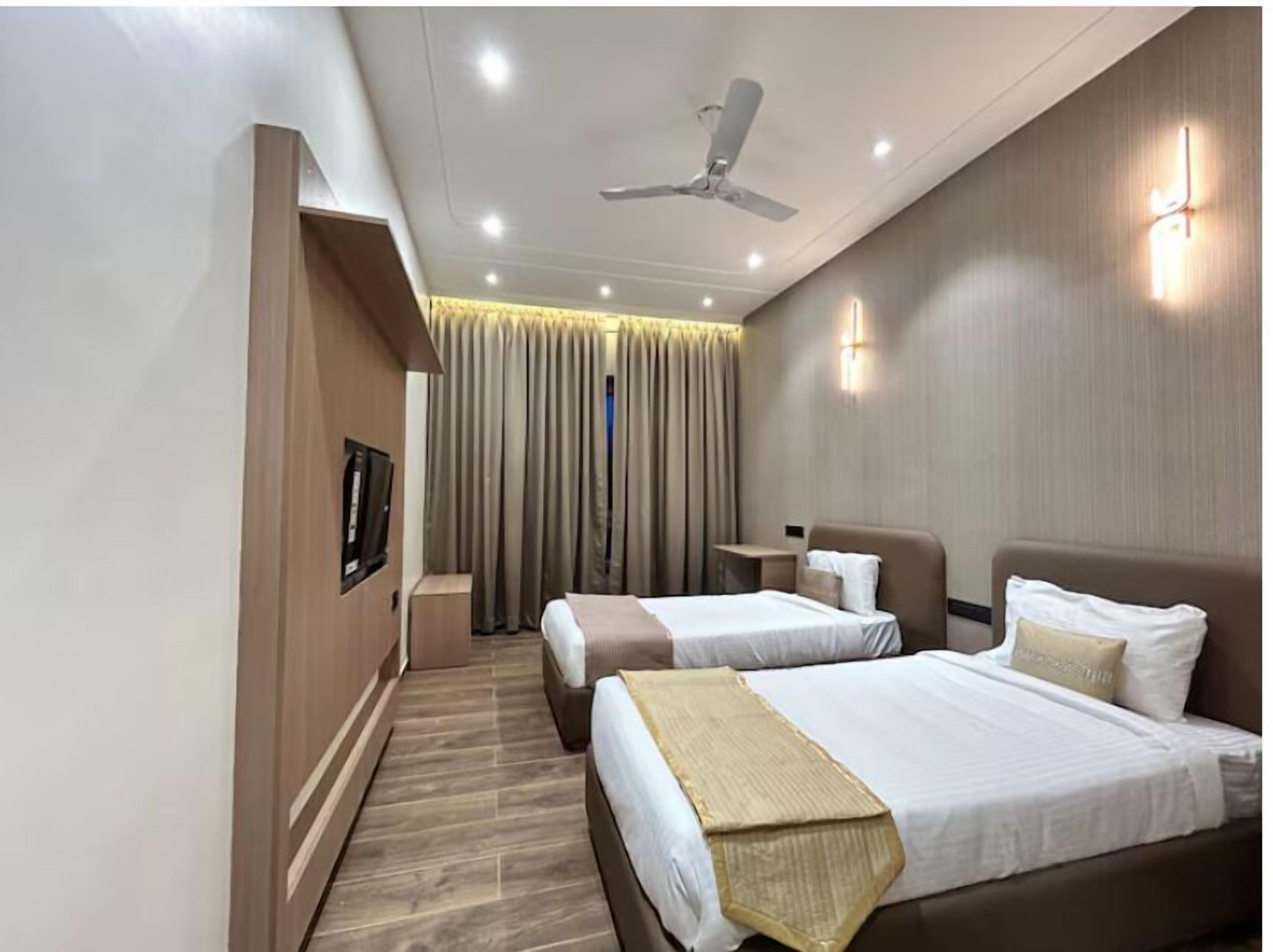 A hotel room with two beds, a ceiling fan, recessed lighting, a TV, and a view of the outside through curtains.