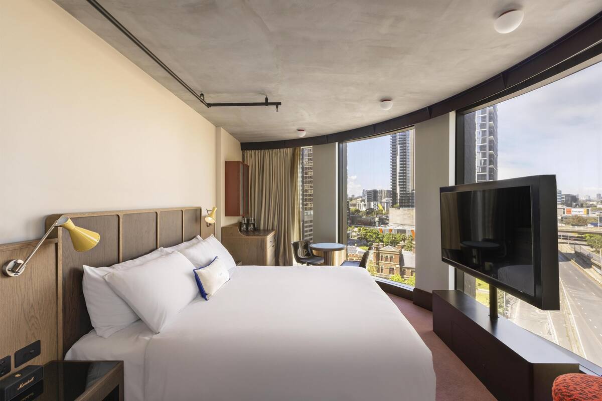 A hotel room with a large bed, a TV, a desk, and a view of the city.