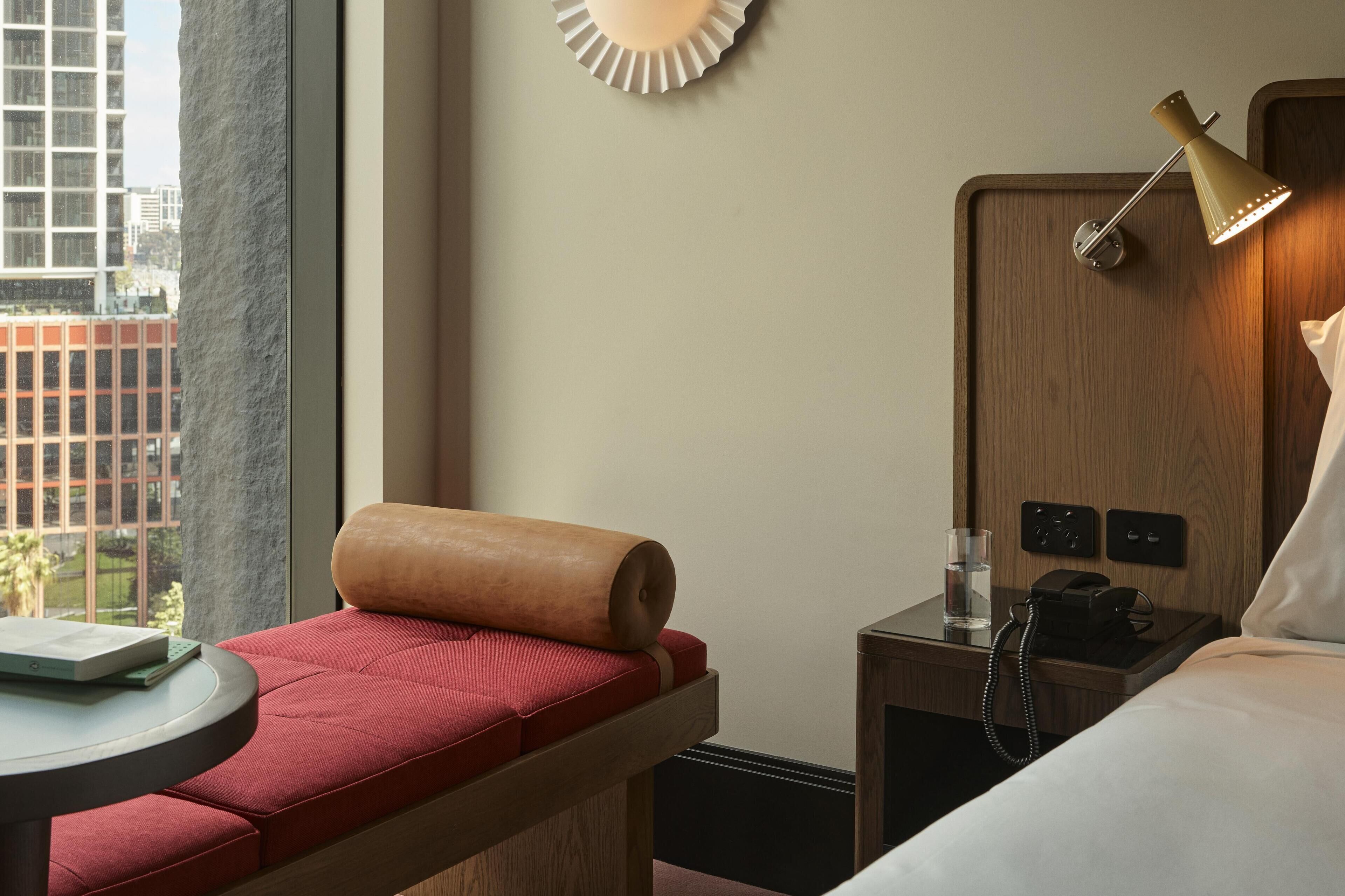 A hotel room with a bed, a bedside table with a lamp, a phone, and a small table with a book and glasses.
