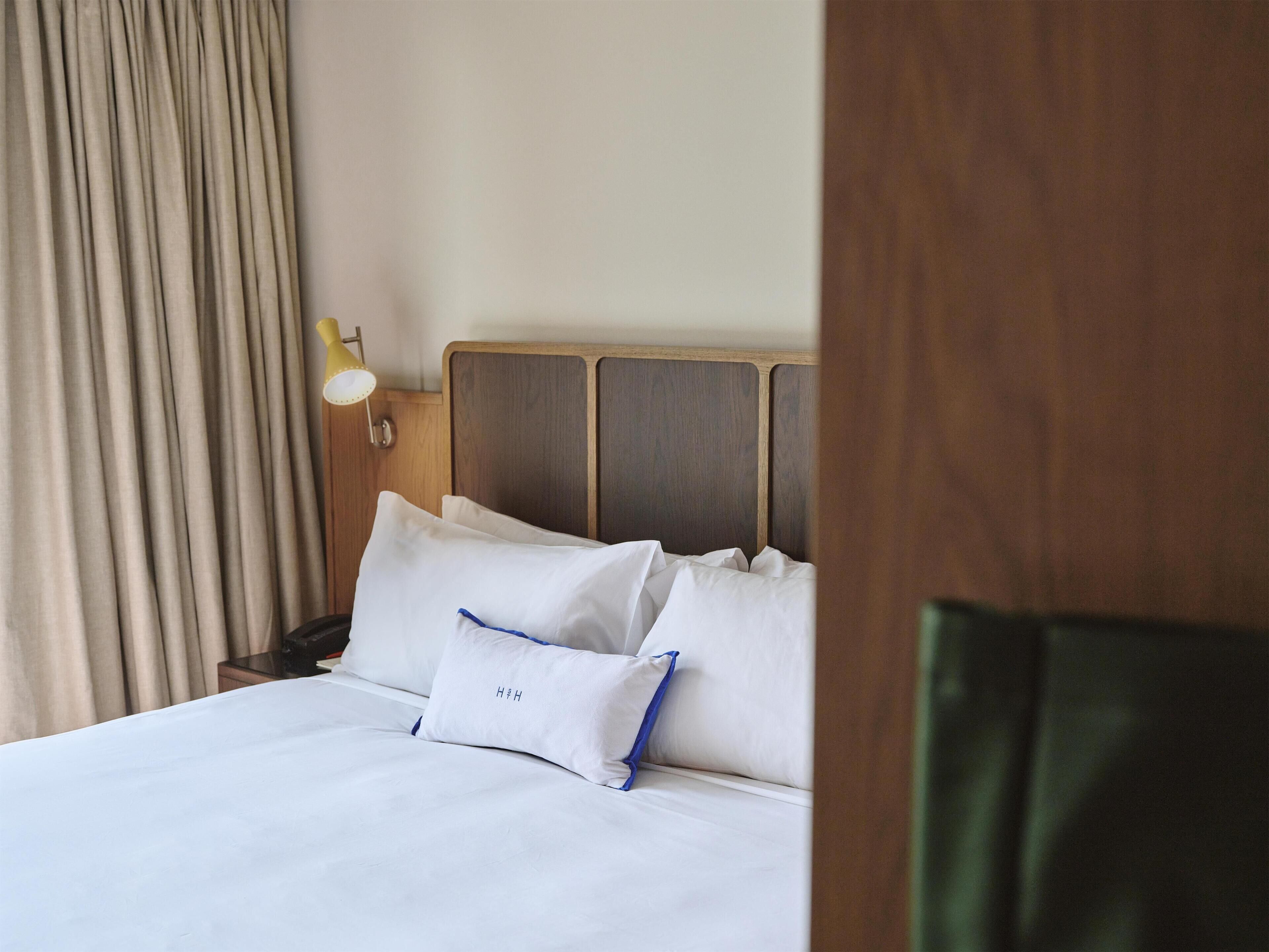 A neatly made bed with white linens and blue accents, a wooden headboard, a bedside lamp, and a curtain partially drawn.