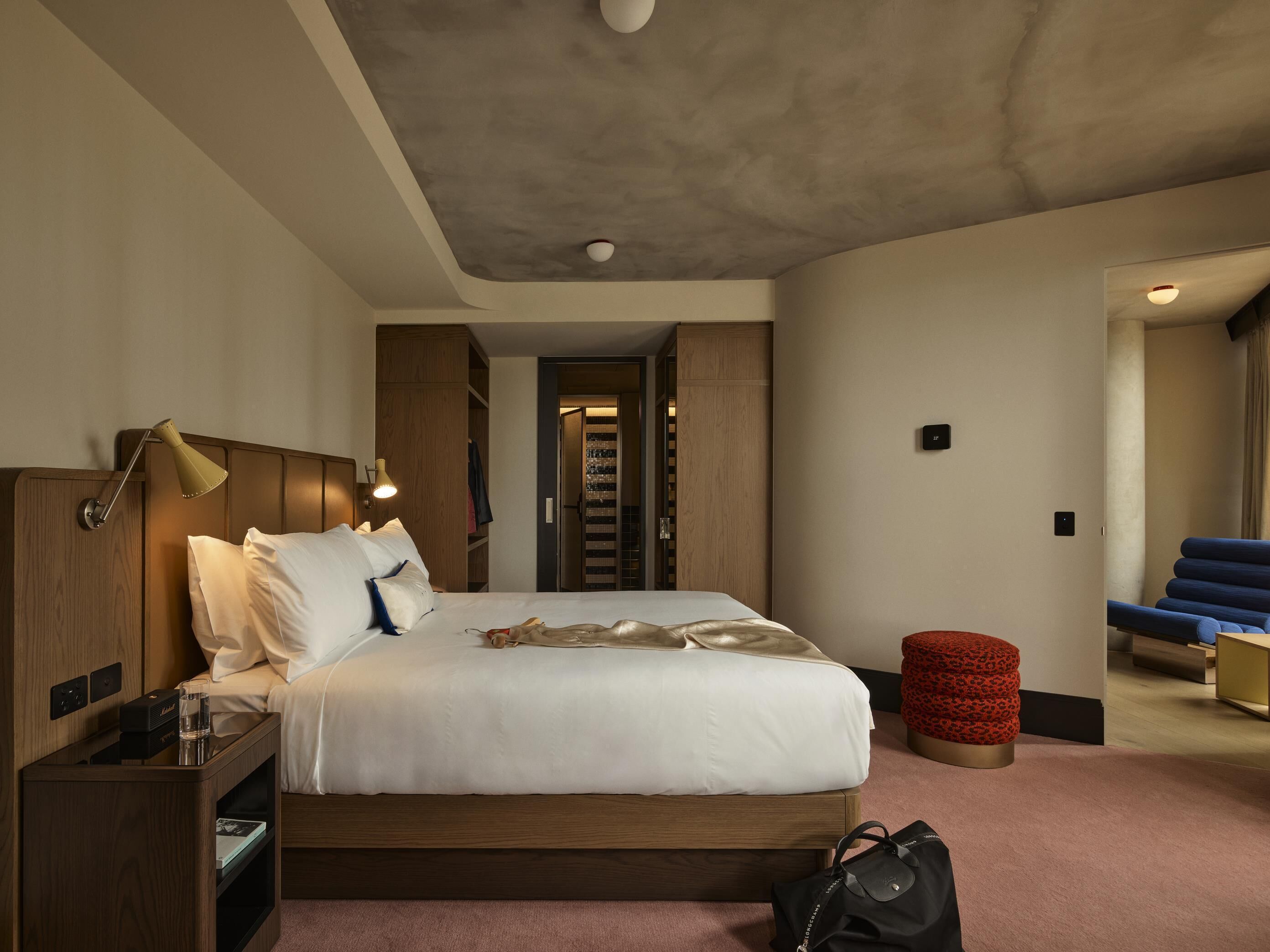 A hotel room with a bed, bedside lamp, nightstand, and a red ottoman.