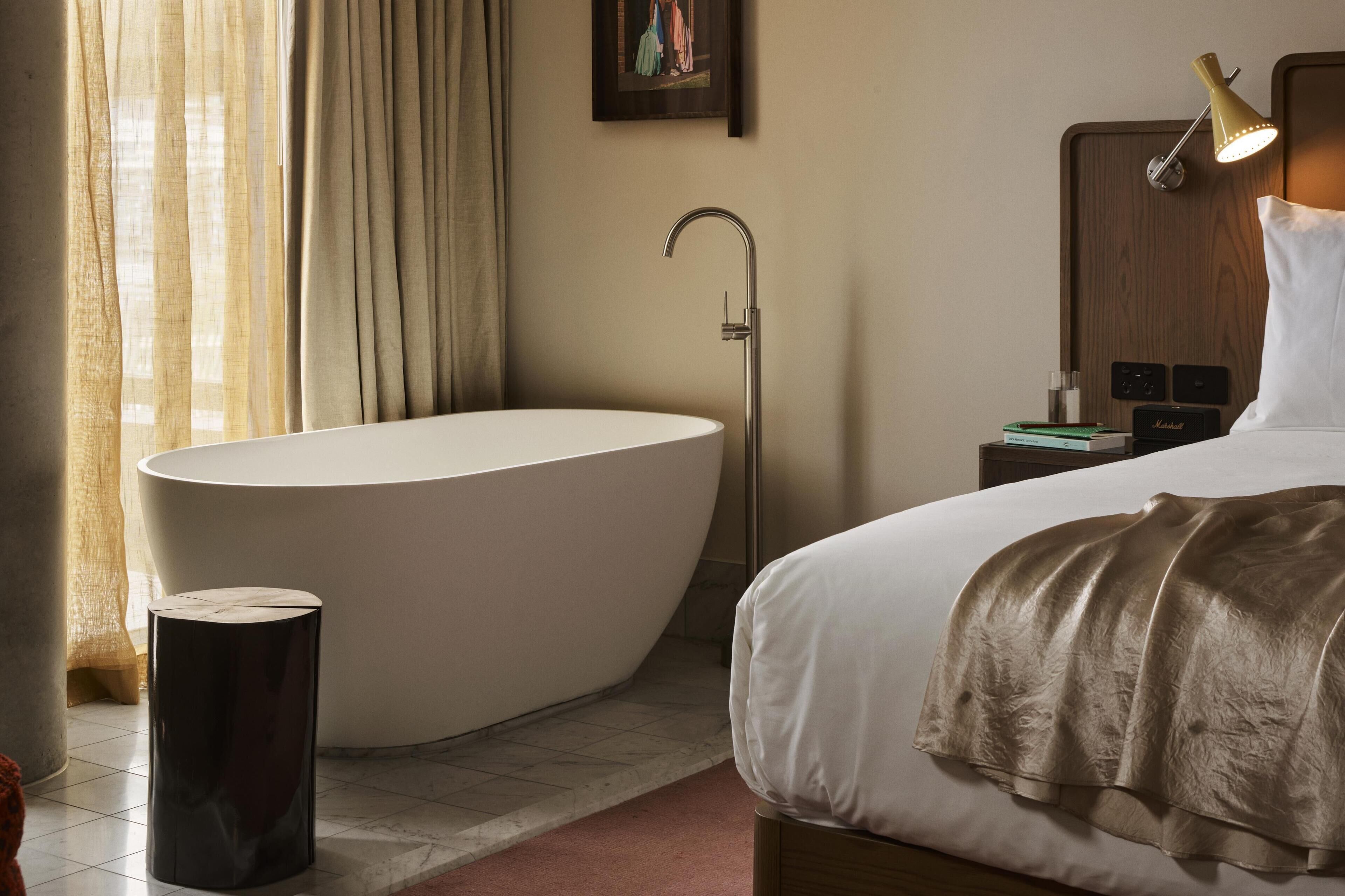 A modern hotel room with a large bathtub, a bedside lamp, a nightstand, and a bed with a white comforter.