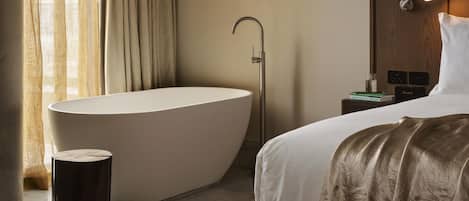 Habitación de hotel moderna con una bañera grande, una lámpara de noche, una mesita de noche y una cama con edredón blanco.