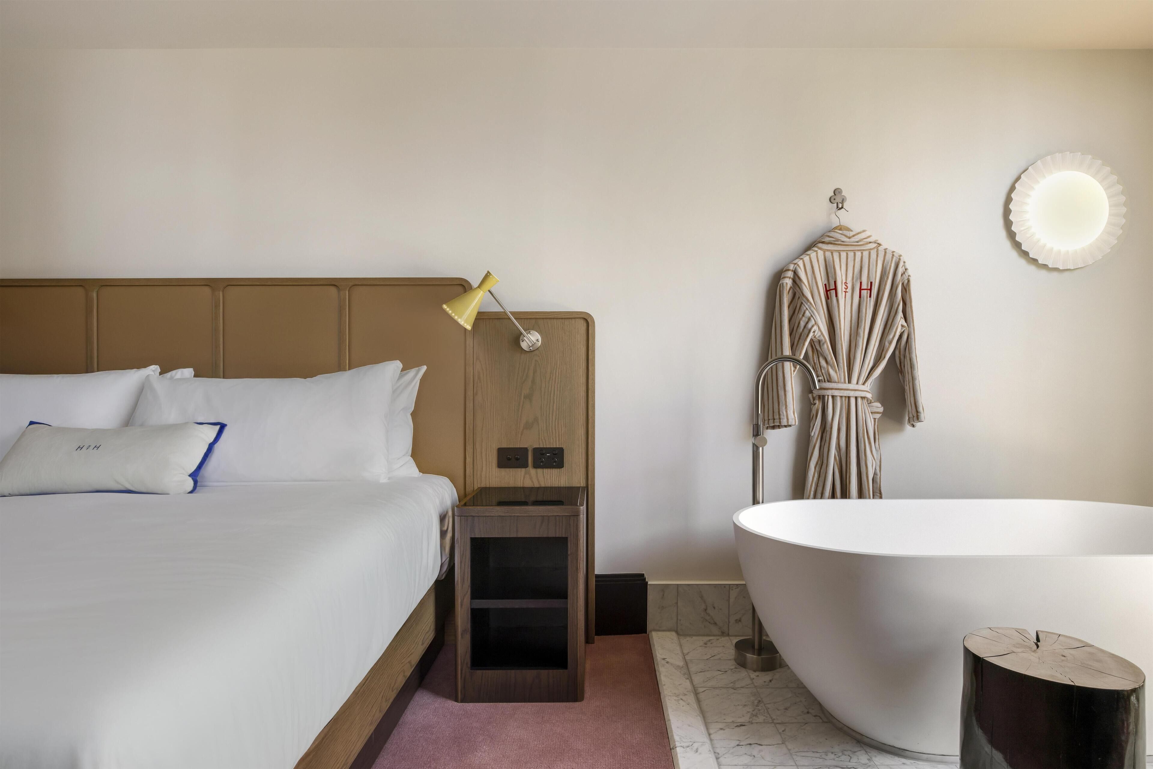 A hotel room with a bed, bedside lamp, robe, and a freestanding bathtub.