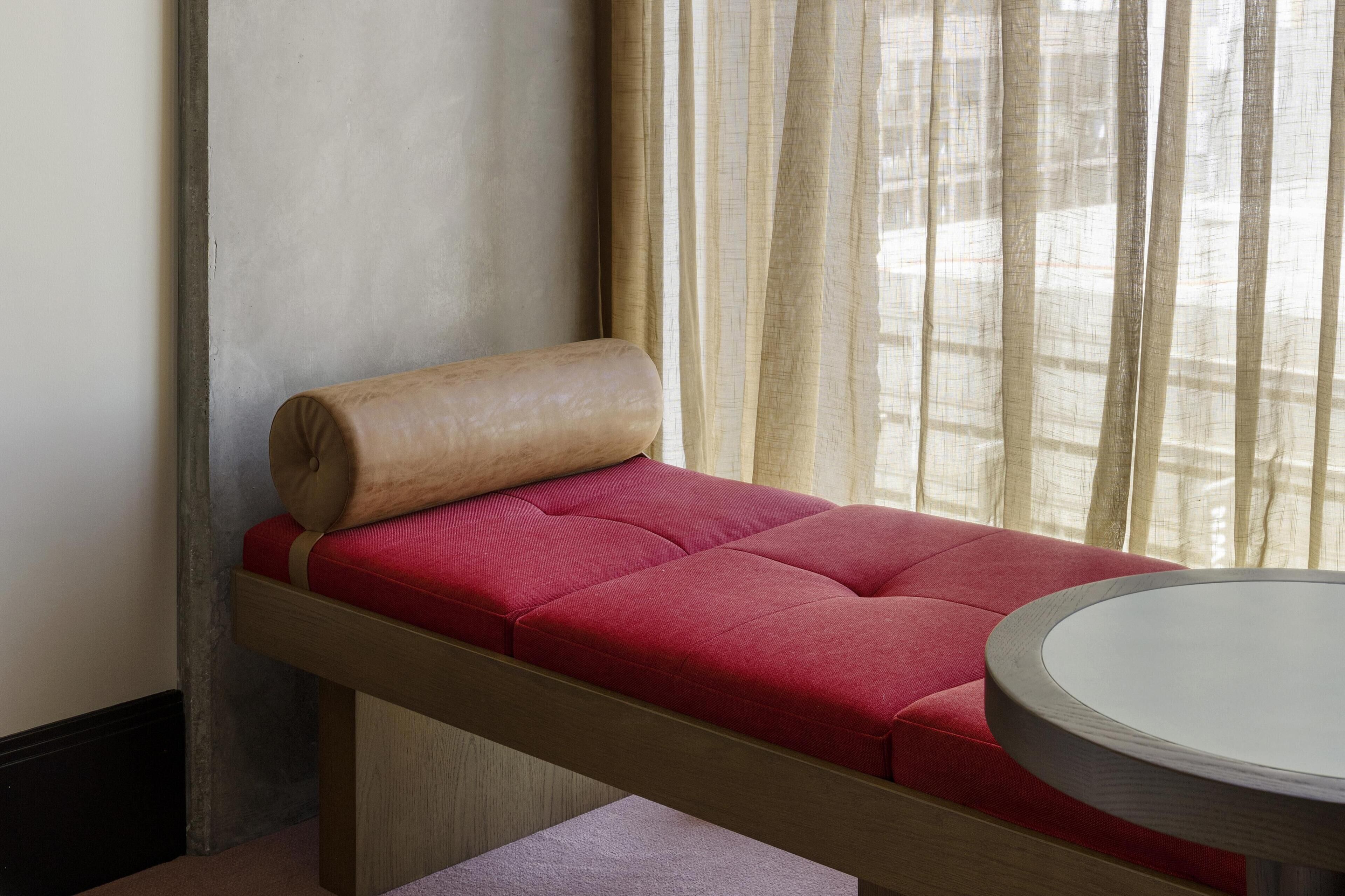 Een kleine kamer met een enkel bed, houten frame, rode matras en een ronde tafel.