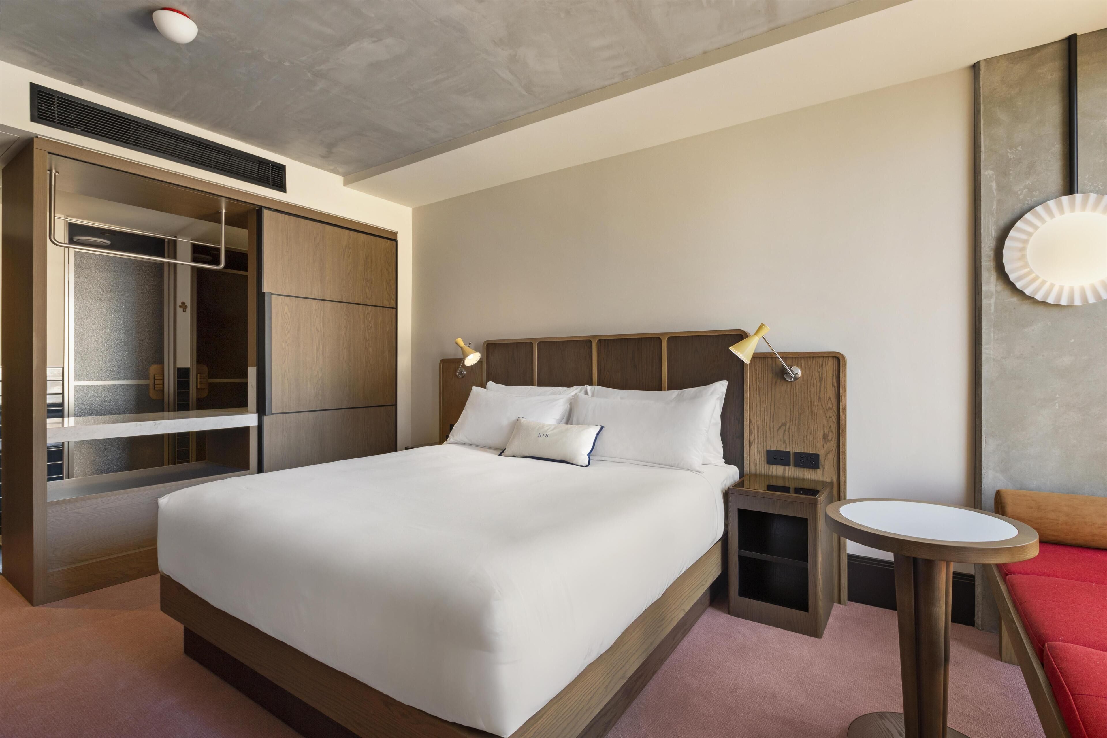 A modern hotel room with a large bed, a wardrobe, a bedside table, and a small round table.