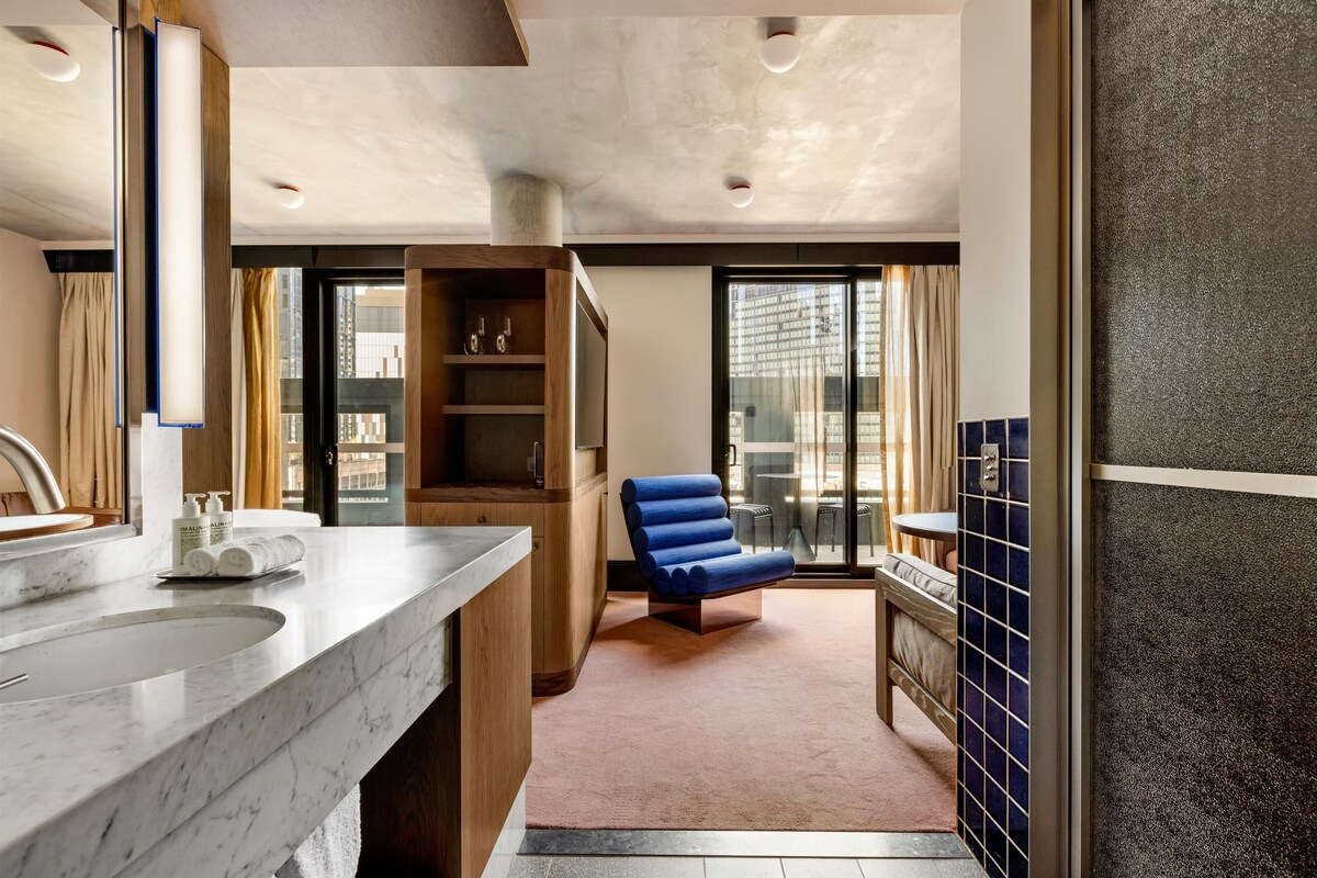 A modern bathroom with a marble sink, wooden cabinets, a blue chair, and a view of the city through large windows.