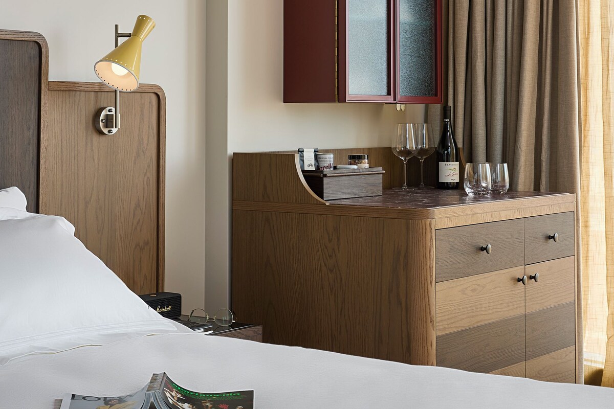 Quarto de hotel com cama, cómoda de madeira, minibar e janela com cortinas.