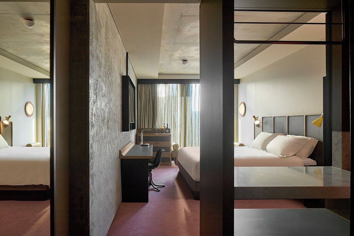 Quarto de hotel com uma cama grande, uma escrivaninha com cadeira, uma mesinha de toucador com espelho e uma janela com cortinas.