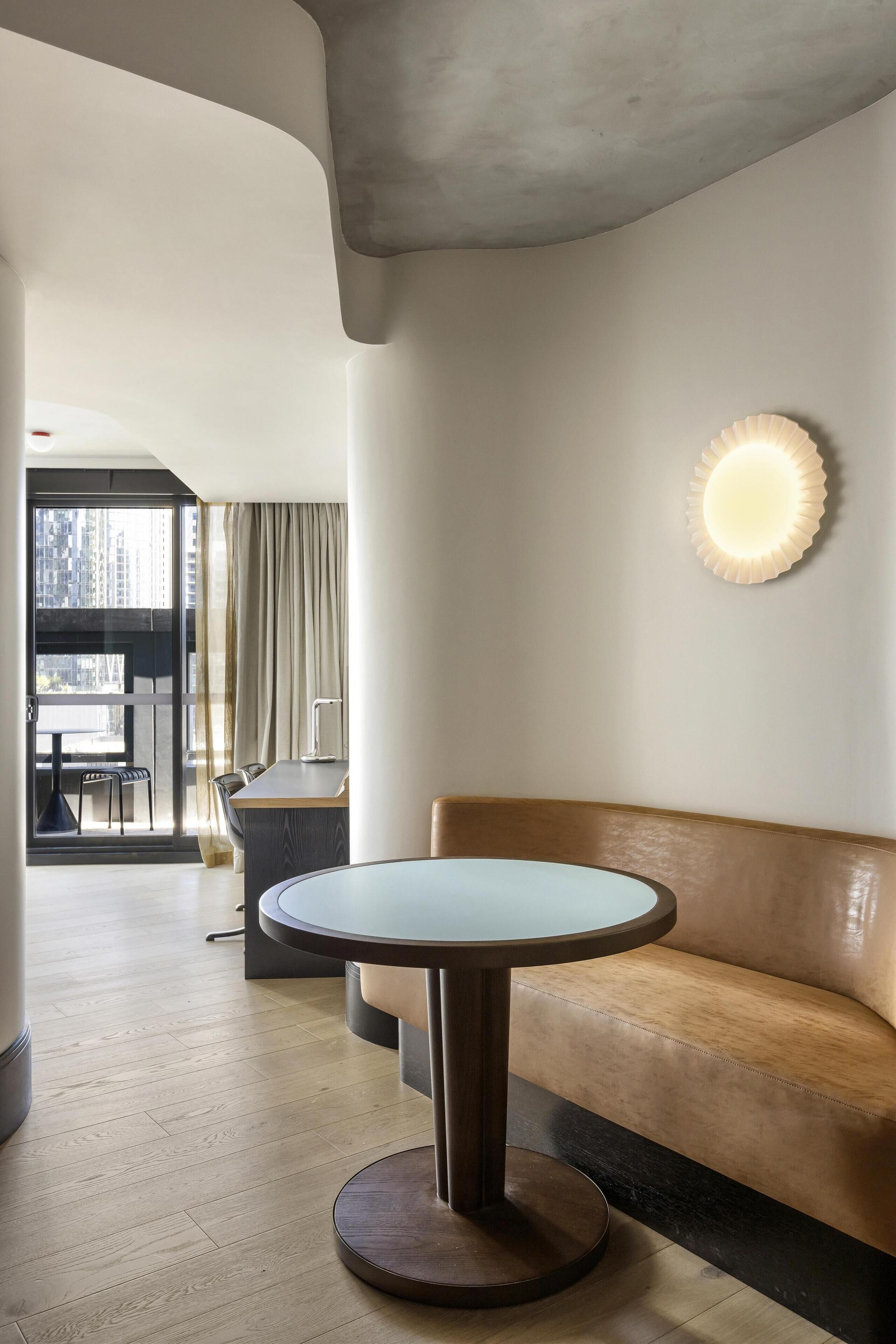 Una habitación de hotel moderna con una mesa redonda, un sofá de cuero, una zona de comedor con una mesa y sillas, y un ventanal con cortinas.
