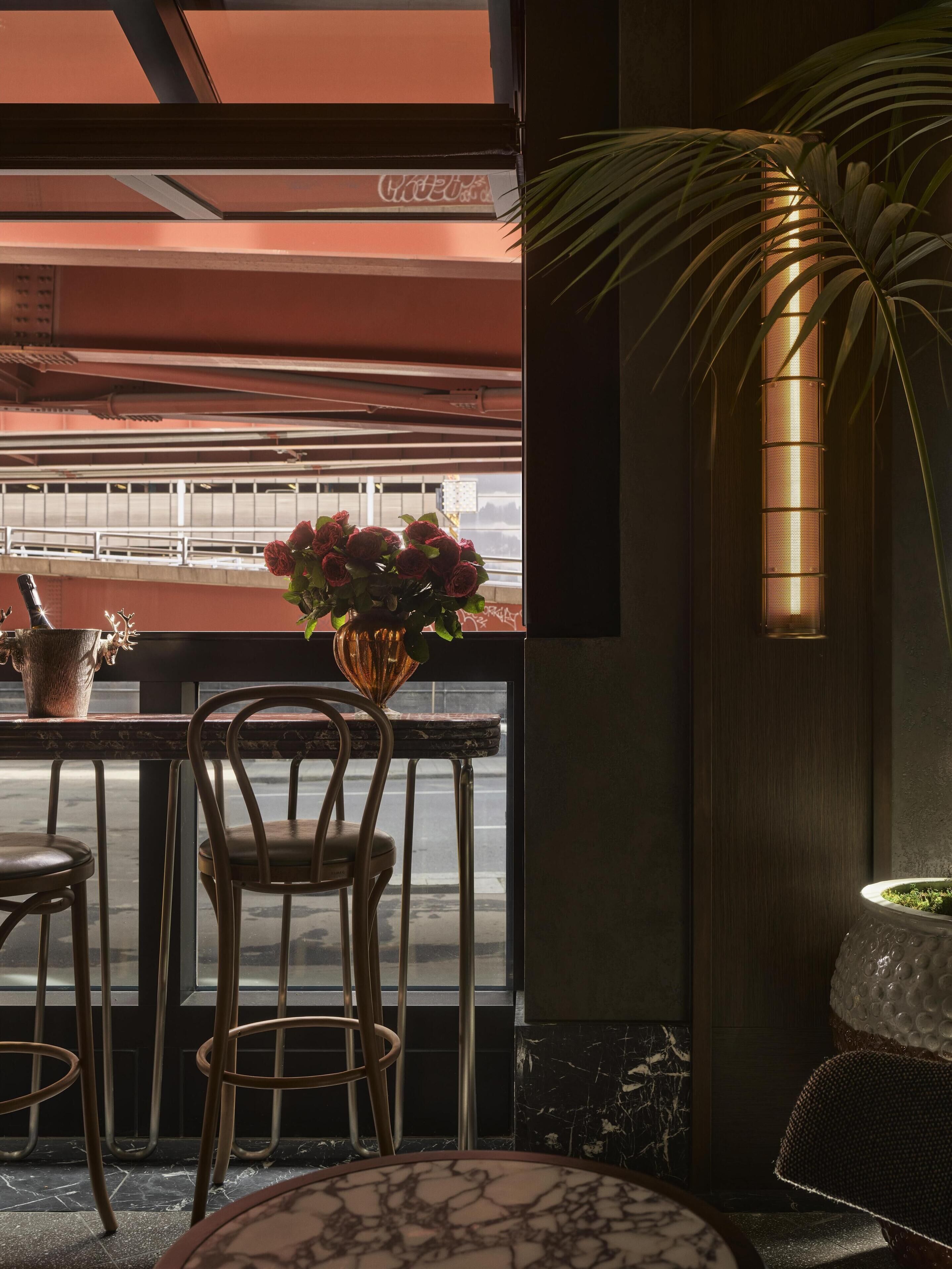 A restaurant interior with a view of a parking garage, a table with chairs, a vase with flowers, and a wall-mounted lamp.