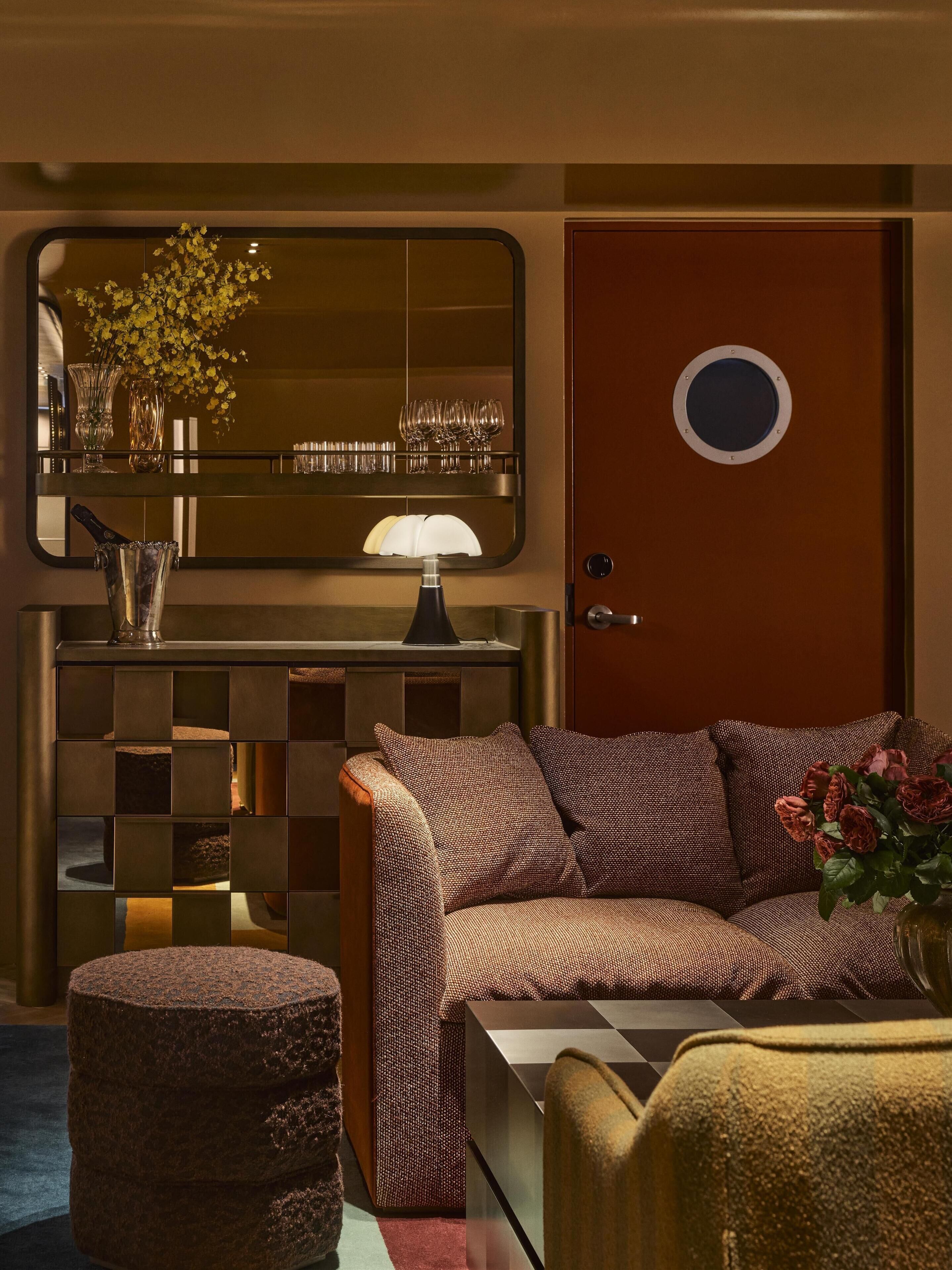 A room with a checkered sofa, a small ottoman, a mirror, a lamp, and a door with a round window.