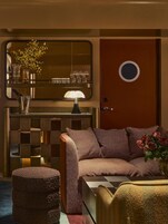 Una habitación con sofá a cuadros, un pequeño otoman, un espejo, una lámpara y una puerta con un ventanal redondo.