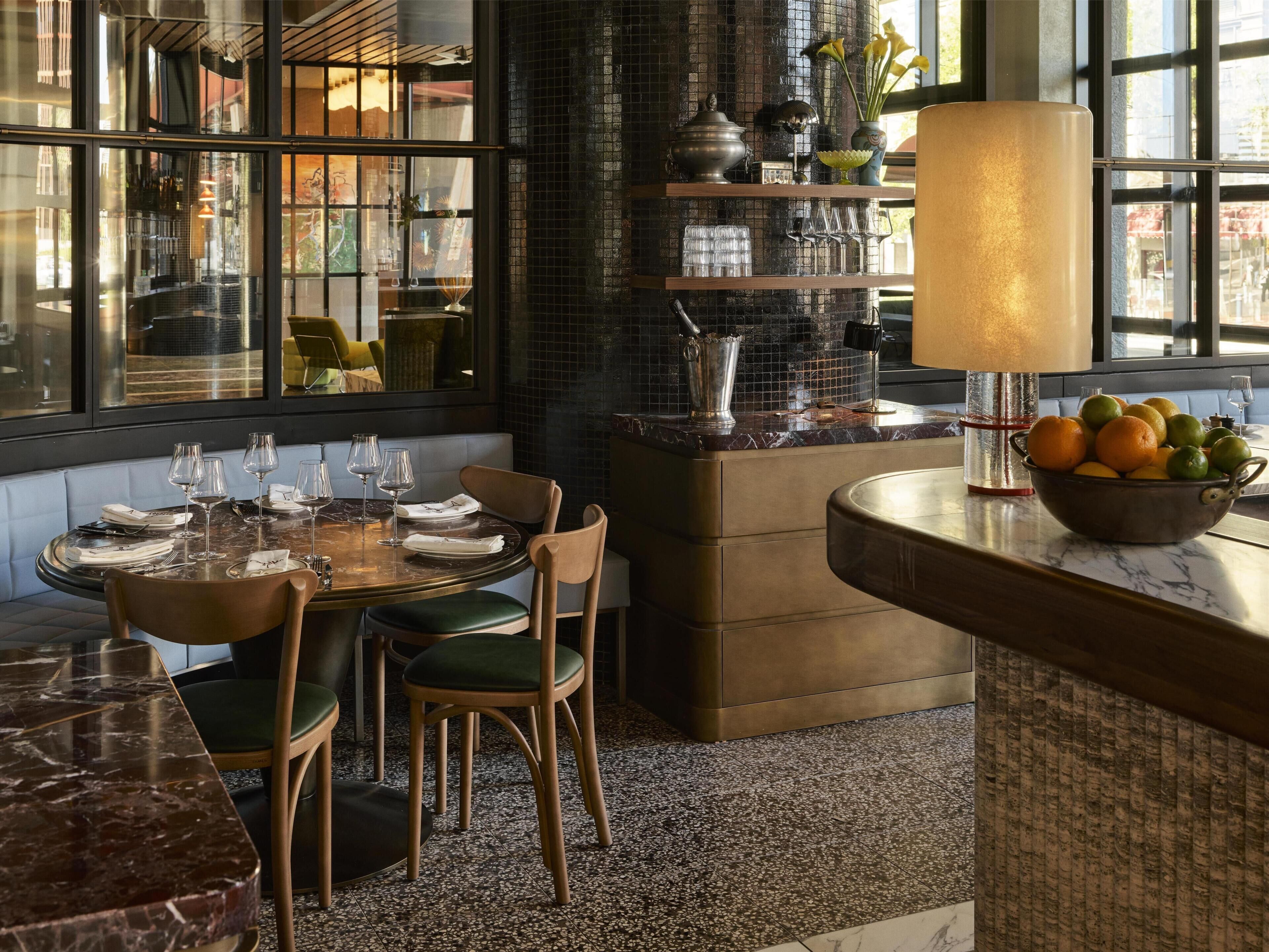 L’intérieur d’un restaurant présente une table dressée pour quatre personnes, un coin bar avec un saladier rempli de fruits et une grande fenêtre offrant une vue sur l’extérieur.