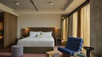 A hotel room with a large bed, a blue armchair, a small ottoman, a bedside table, and a nightstand.