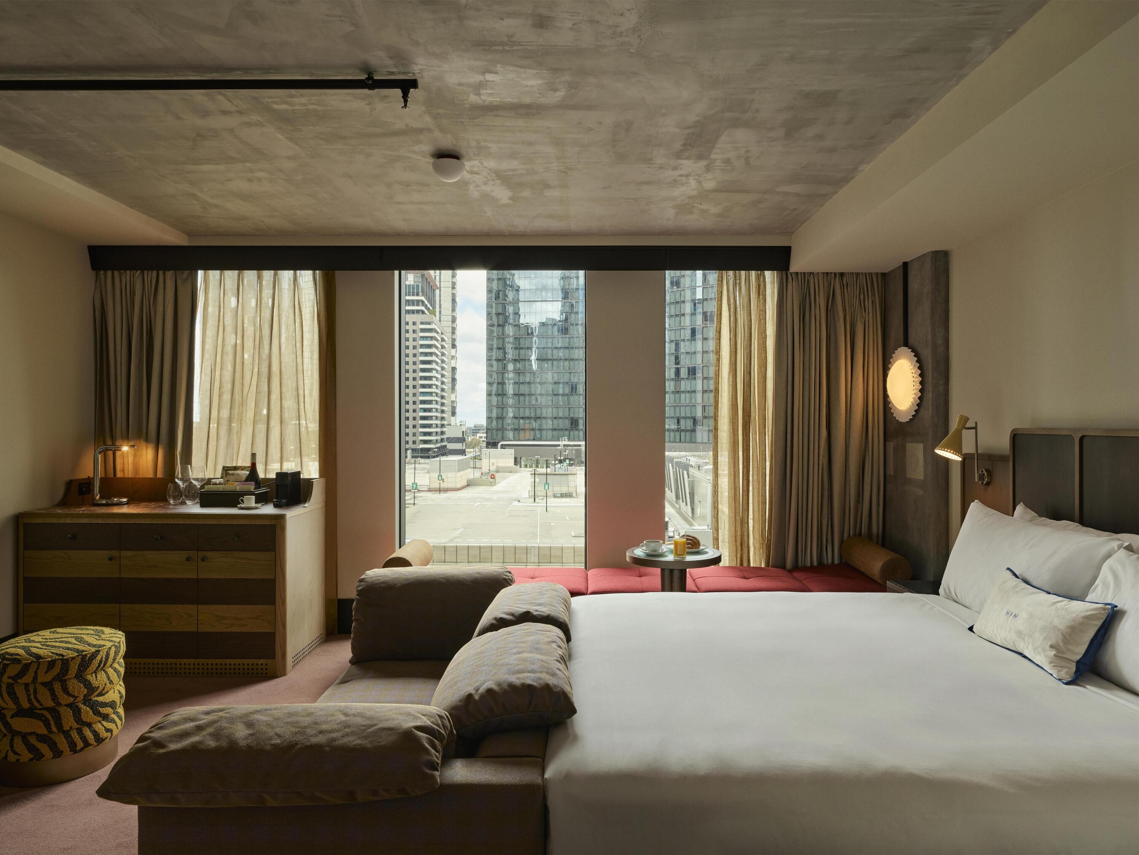 Une chambre d’hôtel avec un grand lit, un bureau avec une lampe, une petite table avec une tasse, et une vue sur le paysage urbain à travers de grandes fenêtres.