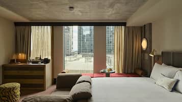 Habitación de hotel con una cama grande, un escritorio con lámpara, una mesita con una taza y vistas a la ciudad a través de amplios ventanales.