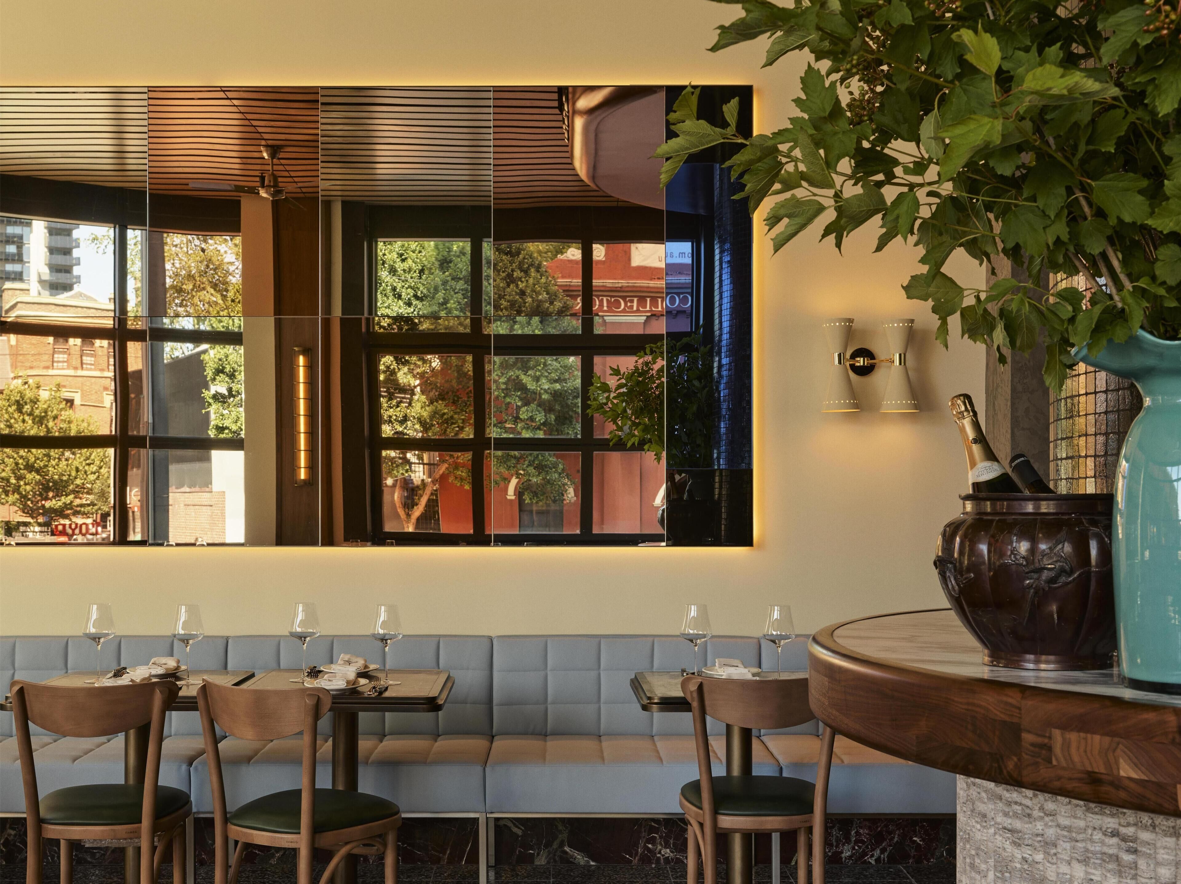 Un restaurante con vistas a un edificio a través de una ventana, una zona de estar con bancos y un florero con una botella sobre una barra.