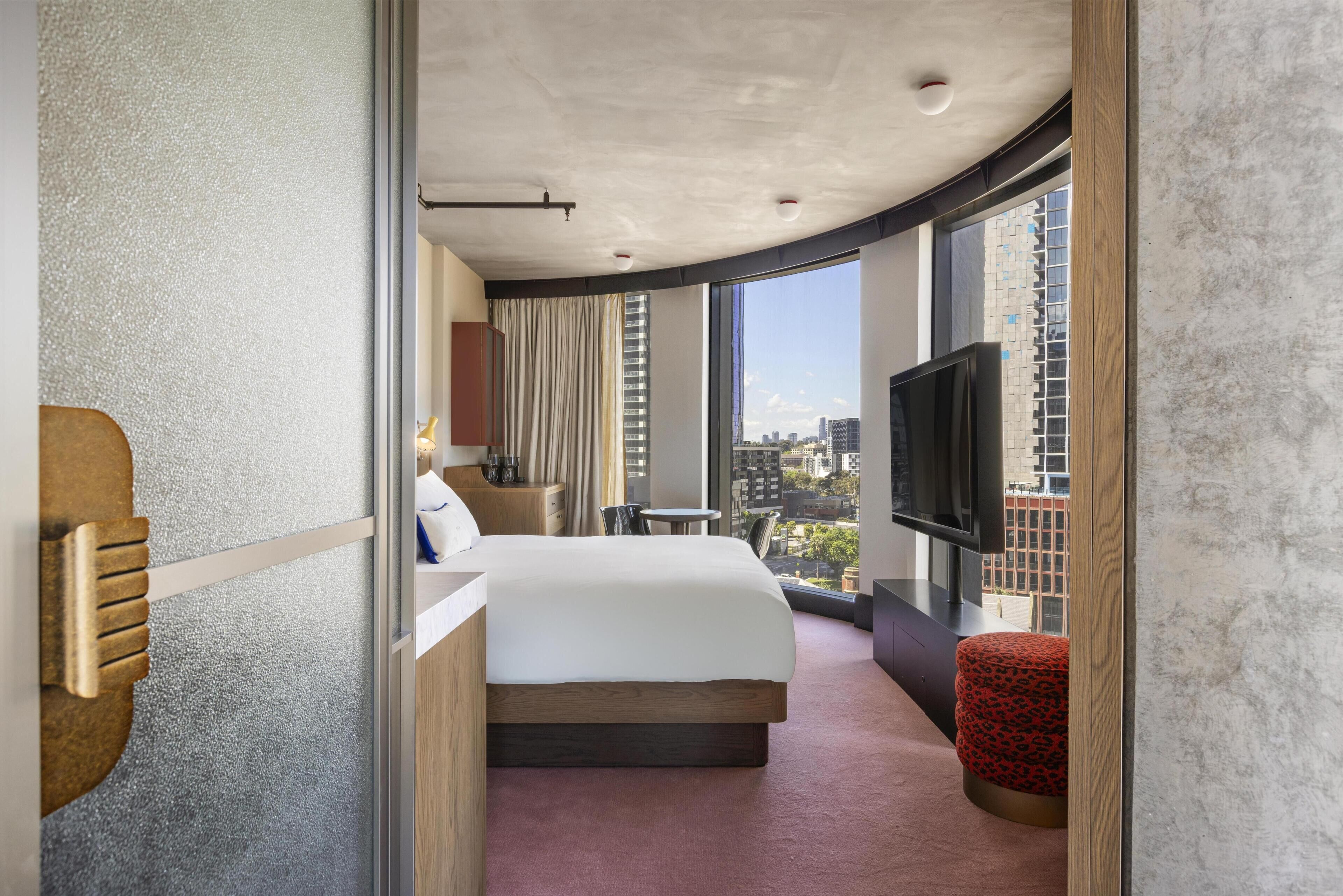 A modern hotel room with a large bed, a TV, a small table, and a view of the city.