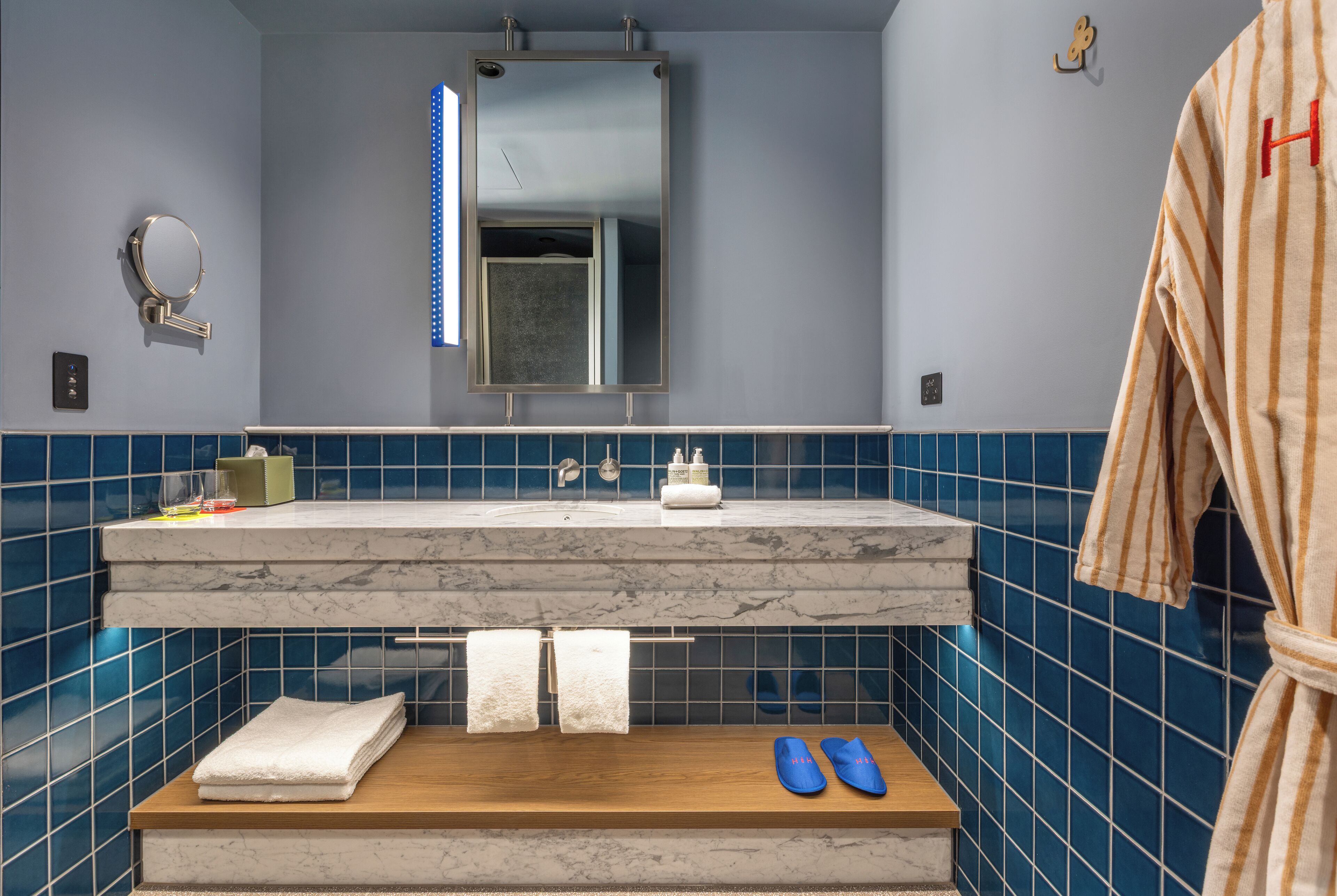 Un baño con encimera de mármol, azulejos azules, un espejo, un perchero para toallas y un par de zapatillas.
