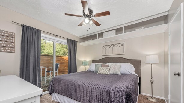 Un dormitorio con cama, ventilador de techo, mesita de noche, lámpara y ventana con cortinas.