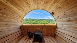 Eine Holzsauna mit Sitzbank und einem Harvia Saunaofen, einem Warnschild und Blick auf einen bewaldeten Hügel.
