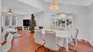 A spacious living area with a large round table, white chairs, a fireplace, a Christmas tree, and a modern chandelier.