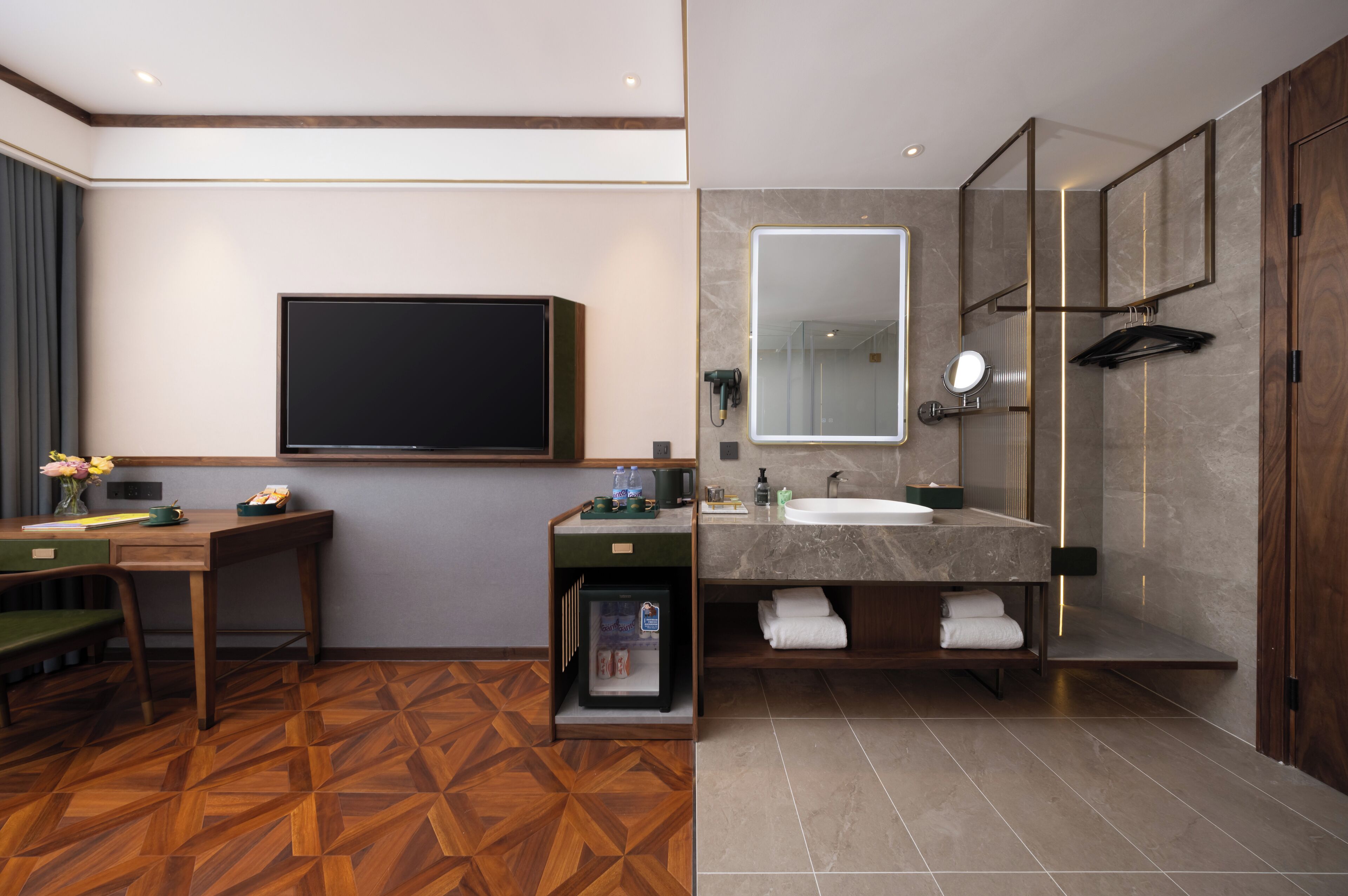 Ein modernes Hotelzimmer mit Flachbildfernseher, Schreibtisch mit Stuhl, Minikühlschrank, großem Spiegel, Waschtisch mit Waschbecken und Duschbereich.