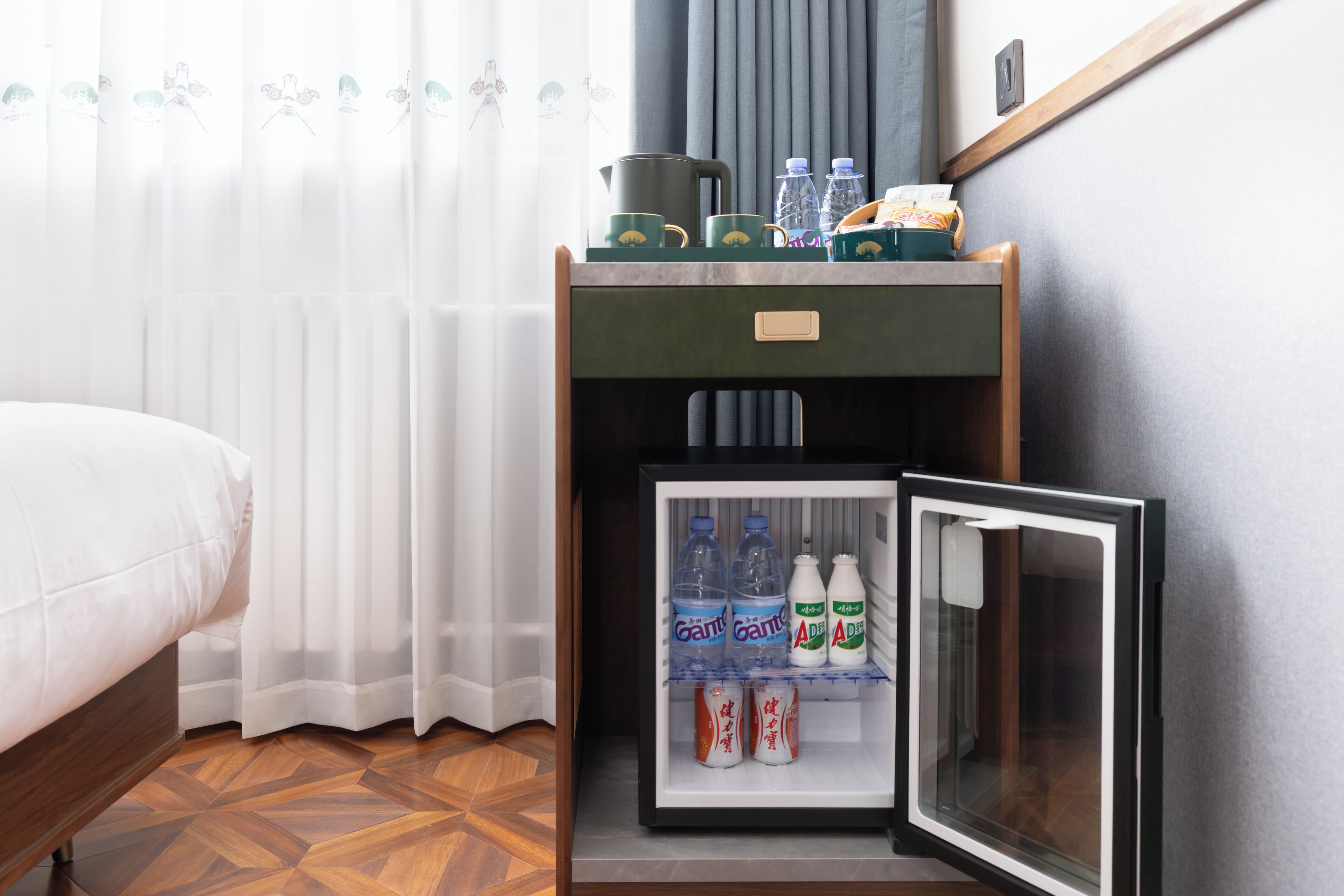 A compact room with a bed, a small kitchenette with a refrigerator stocked with beverages, and a window with sheer curtains.