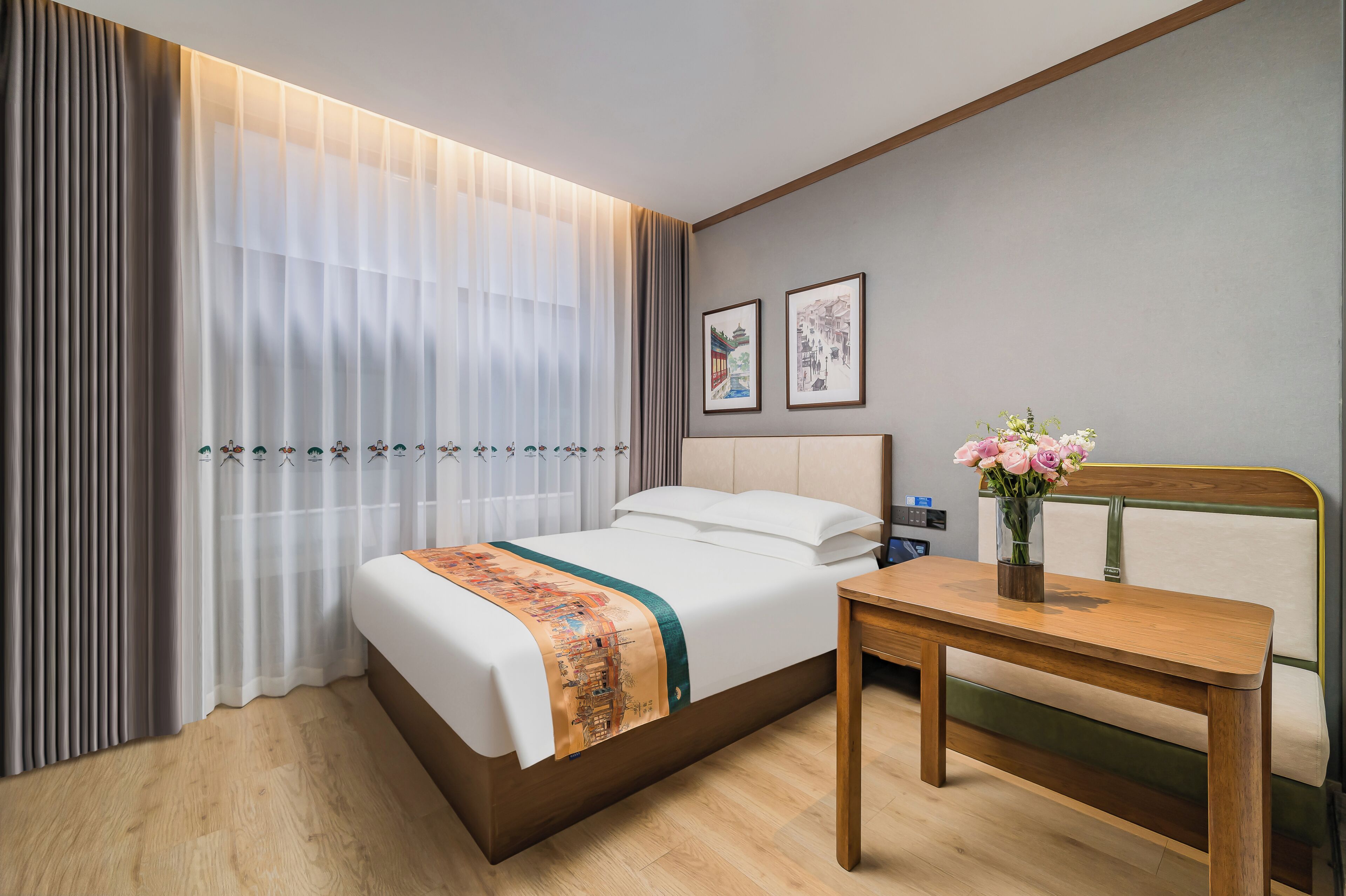 A hotel room with a bed, a wooden table, a vase with flowers, and two framed pictures on the wall.