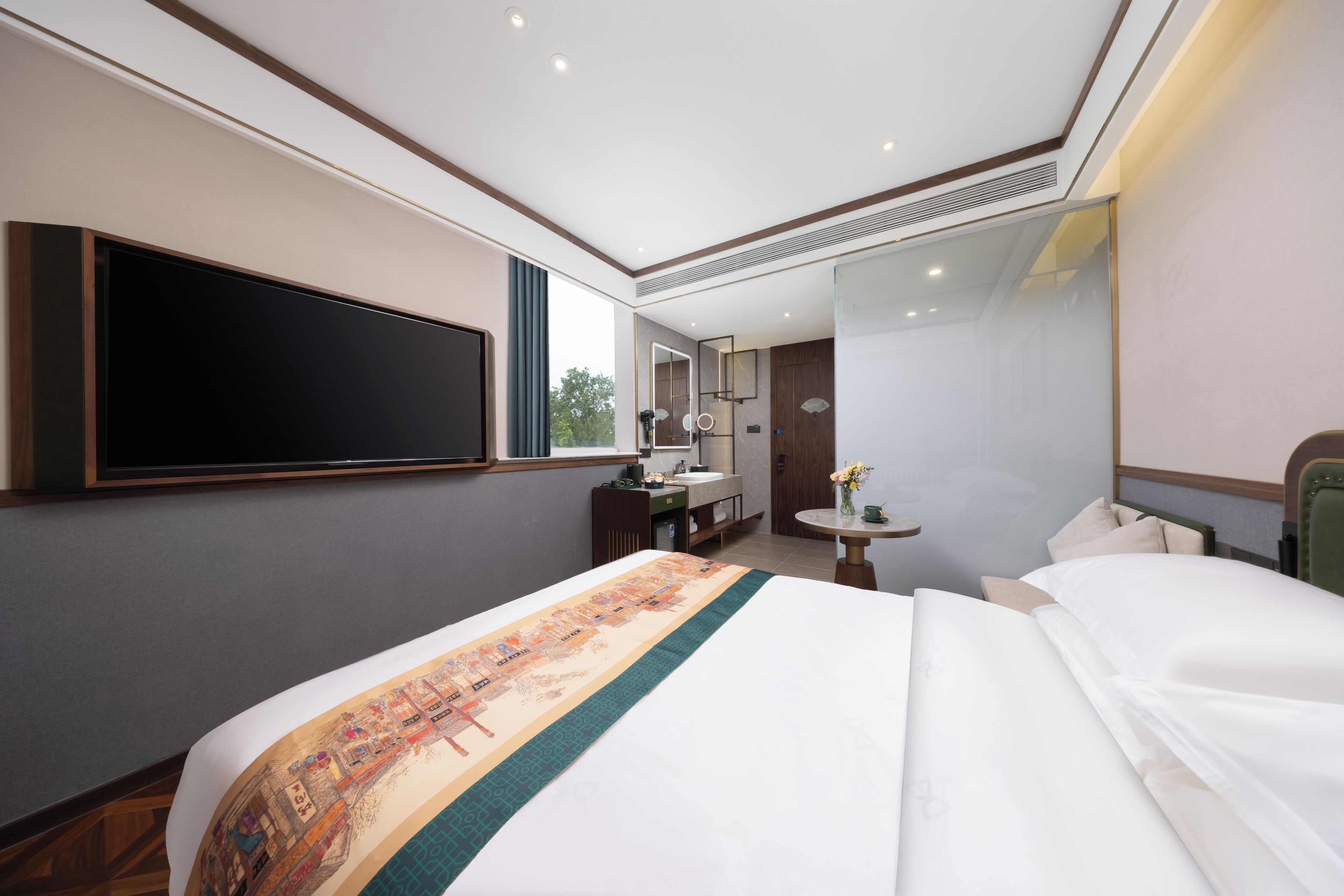 A hotel room with a large bed, a flat-screen TV, a desk with a chair, a round table with a vase, and a large mirror.