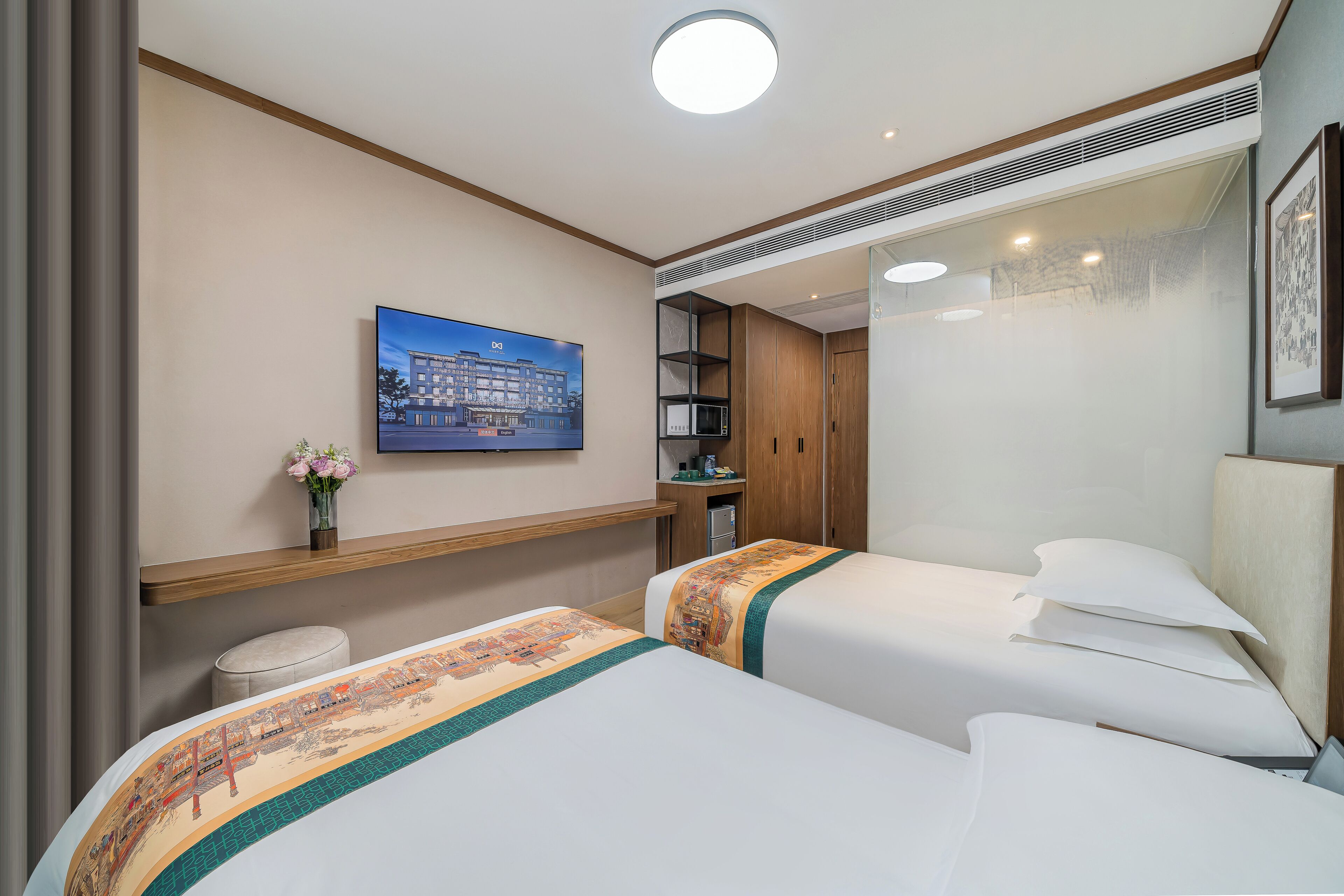A hotel room with two beds, a small stool, a shelf with a vase of flowers, a painting on the wall, and a microwave in the corner.