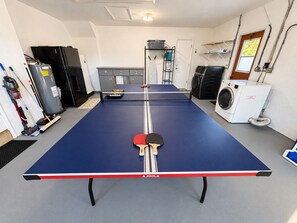 Una mesa de ping pong con mantel rojo y blanco, una paleta de tenis de mesa y un porta pelotas en una habitación con lavadora, calefactor y cesto de ropa.