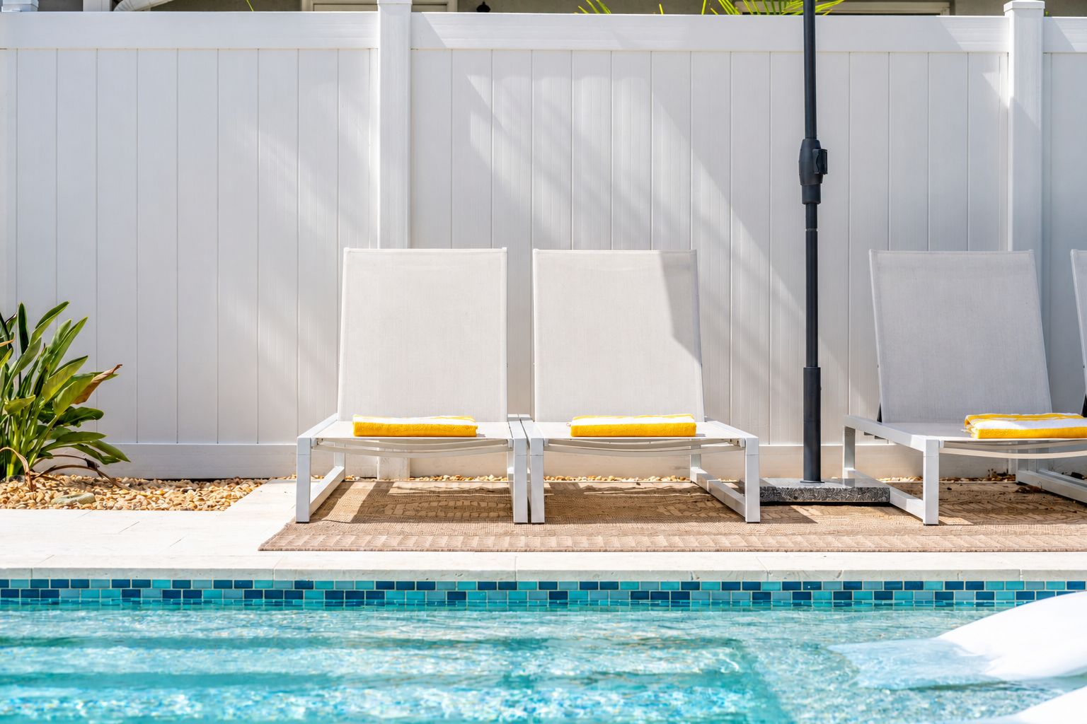 Un espace aménagé au bord de la piscine, avec deux chaises longues blanches et une petite table, une clôture blanche et un bord de piscine recouvert de carreaux.