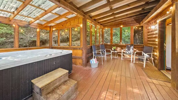A wooden deck with a hot tub, chairs, and a glass-enclosed area with string lights. - Cozy Cabin w/ Hot Tub – Sleeps 8 Comfortably (Pigeon Forge)