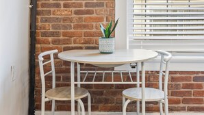 A room with a brick wall, a white door, a window with blinds, a round table with two white chairs, and a potted plant. - Updated Studio in Downtown Long Beach - Walk to Everything (Long Beach)
