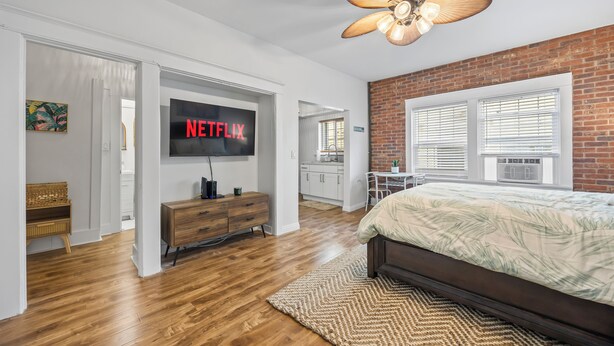 A modern bedroom with a wooden floor, a large bed, a TV displaying 'NETFLIX', a ceiling fan, and a brick wall. - Updated Studio in Downtown Long Beach - Walk to Everything (Long Beach)