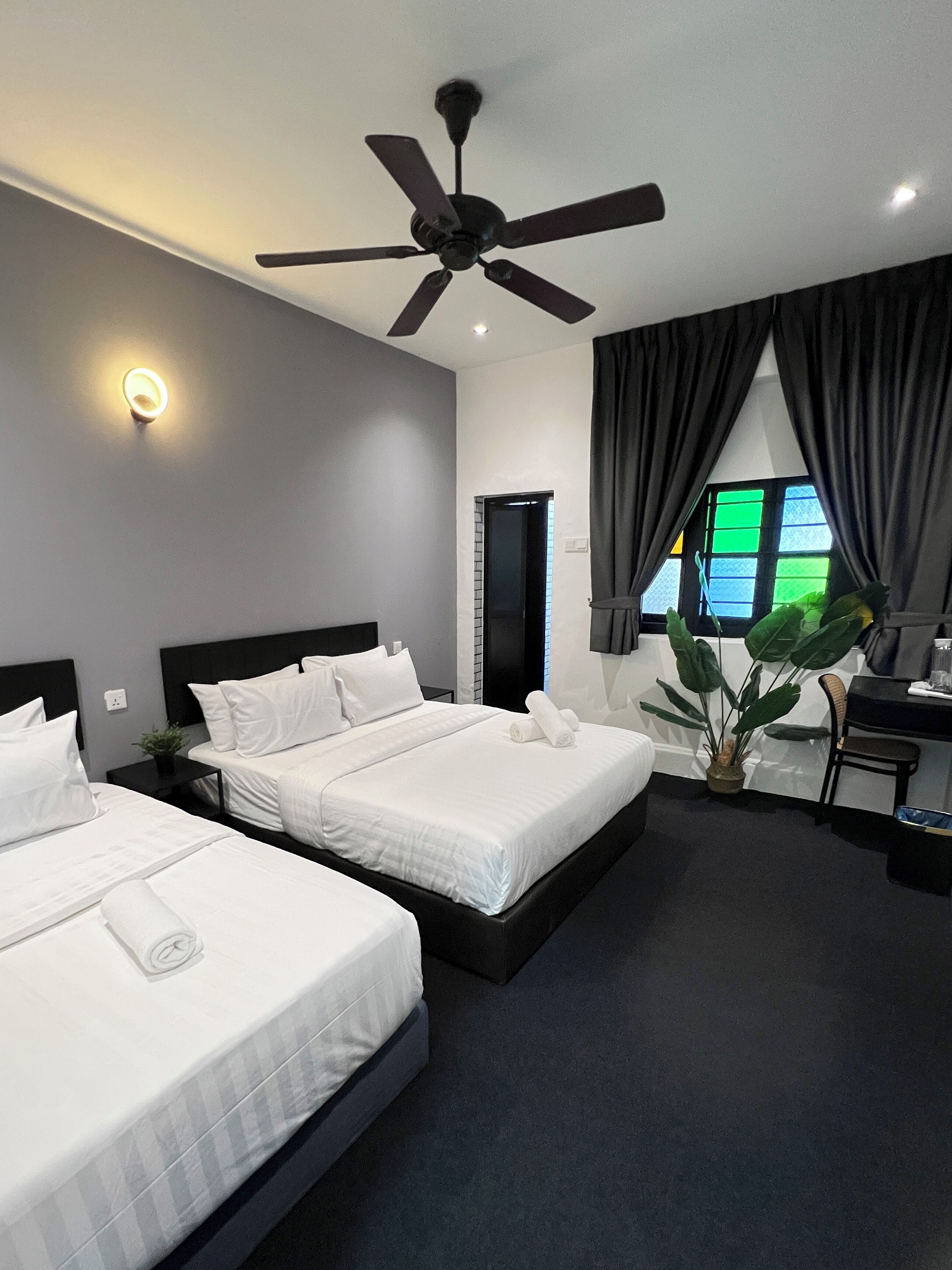 A modern hotel room with two beds, a ceiling fan, a large window with curtains, and a small desk with a chair.