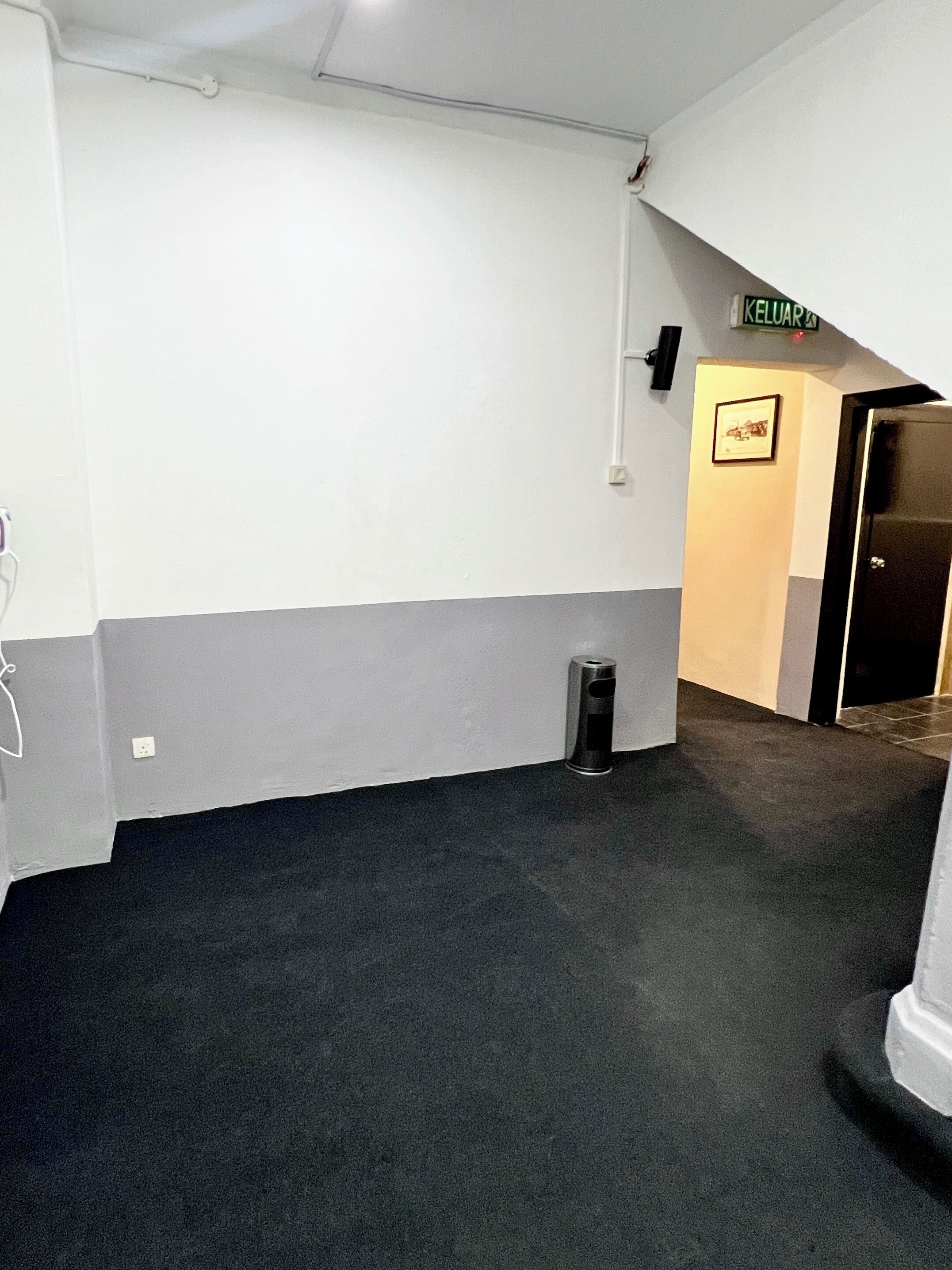 A hallway with a grey and white wall, a black trash bin, and a door with a sign above it.